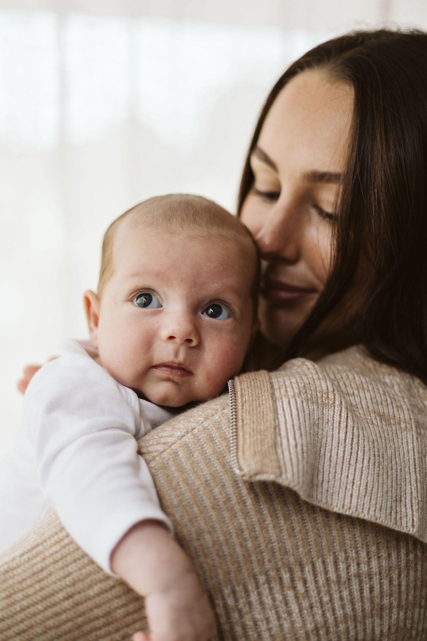 Mother tenderly holds newborn baby with blue eyes close to her cheek.
