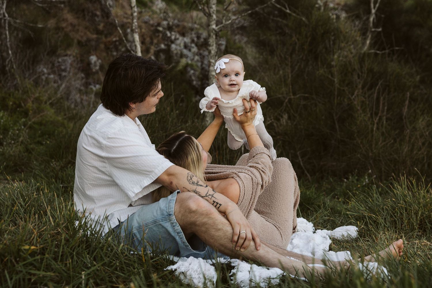 Two women sitting on grass in a forest, joyfully holding a baby girl in a white dress.