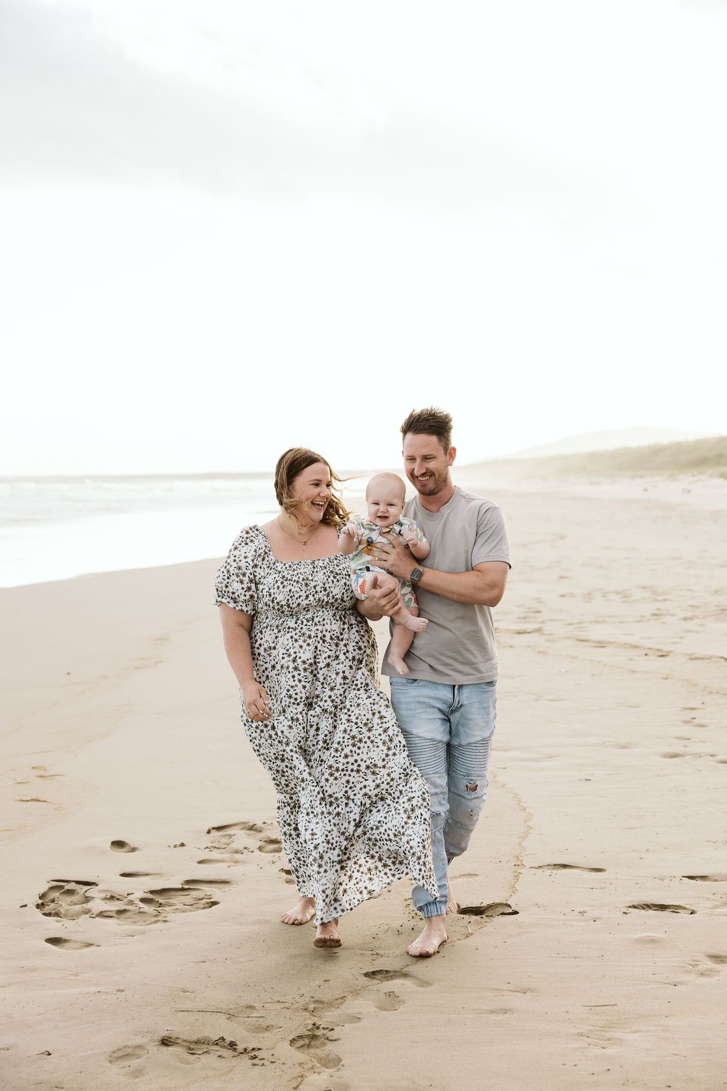 Happy family with baby strolling on sandy beach, parents smiling, casual attire, overcast sky, ocean waves in background.