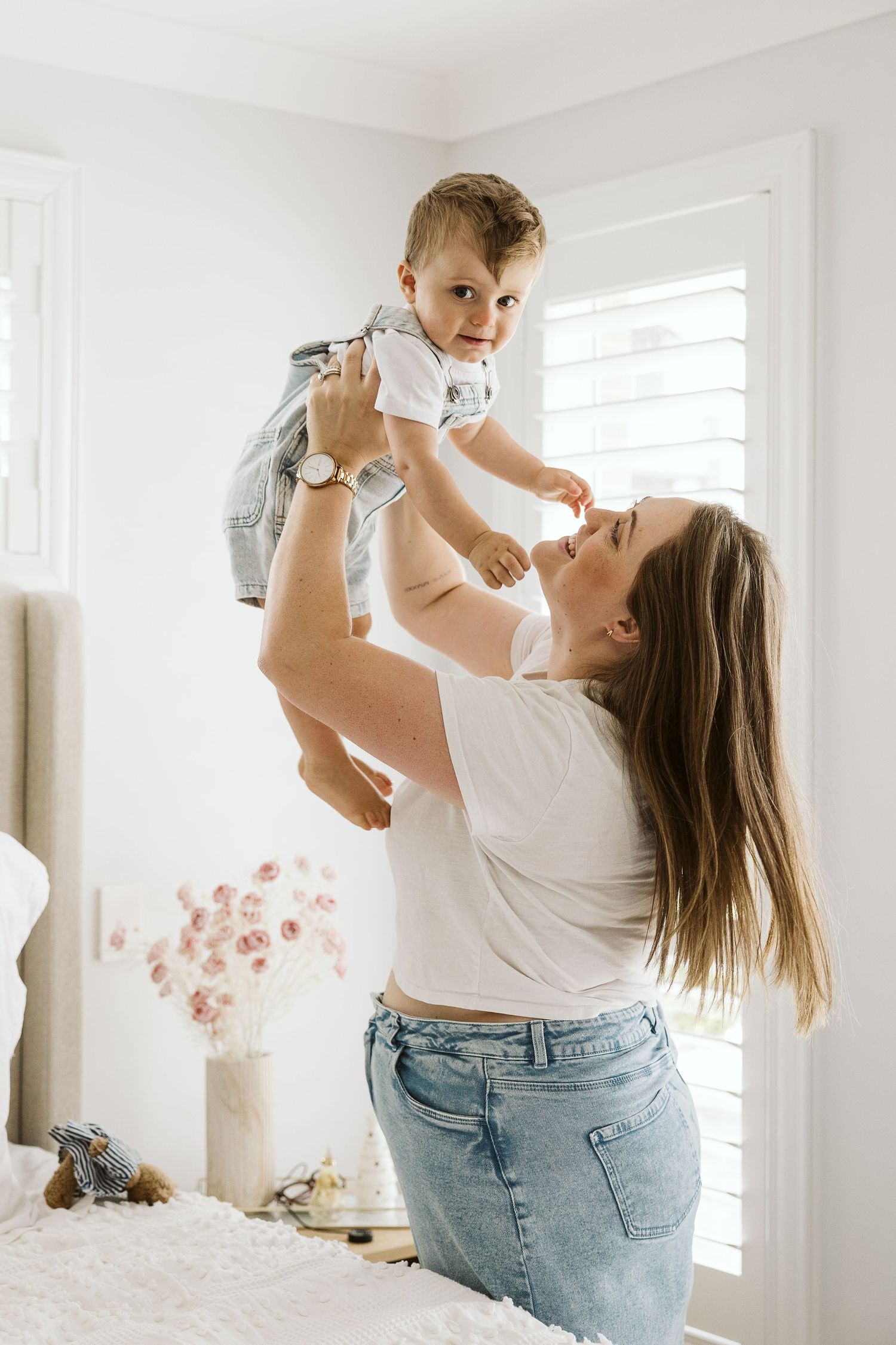 Joyful mother lifting smiling baby boy in denim overalls in bright white bedroom.