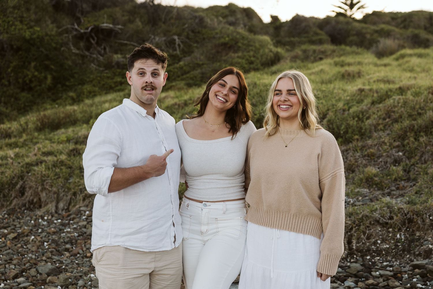 Three young adults smiling outdoors near coastal greenery, two women and a man in casual neutral outfits.
