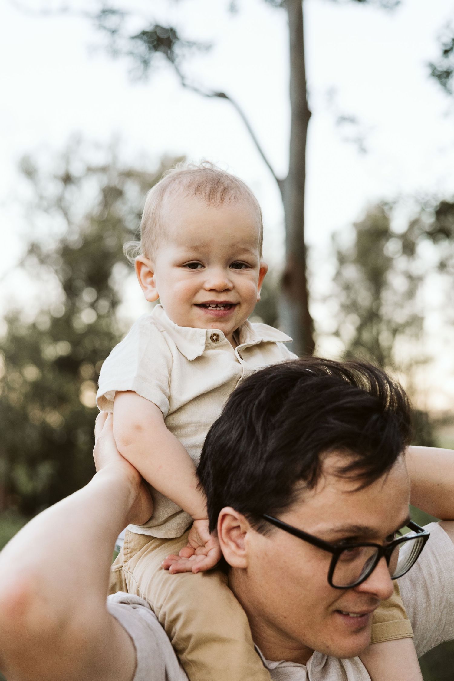 A smiling baby sits on a man's shoulders outdoors, enjoying a piggyback ride in a sunny park setting.