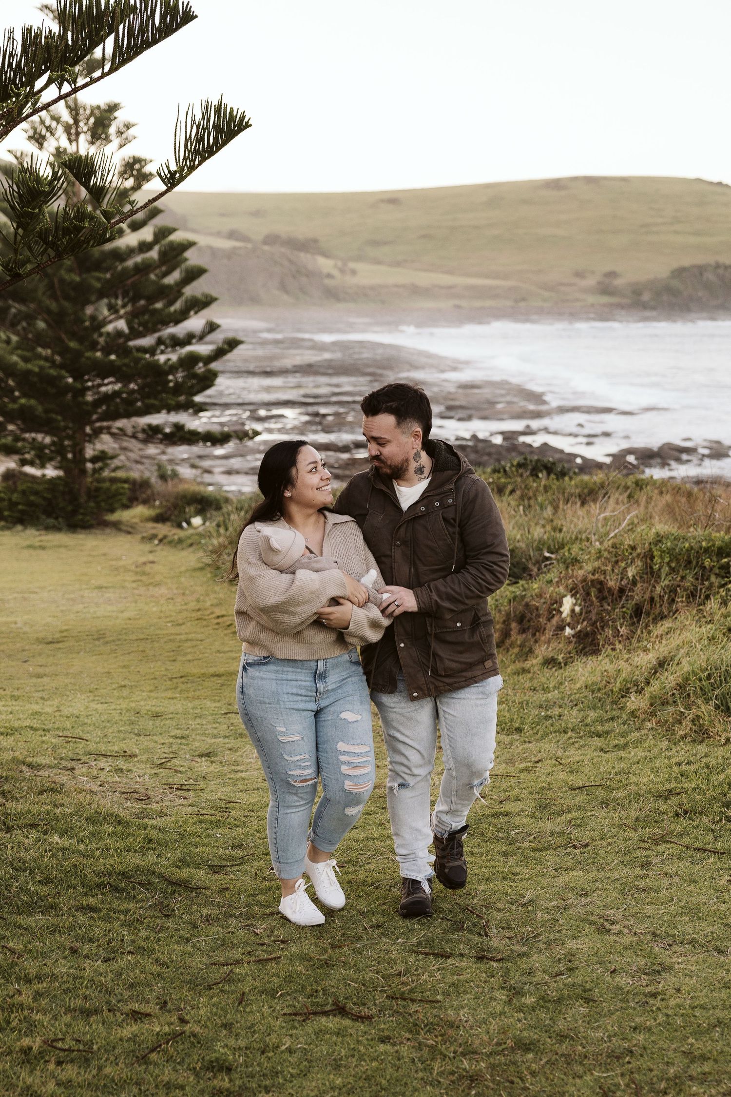 Happy couple walking together on green coastal hillside, smiling at each other near the ocean.