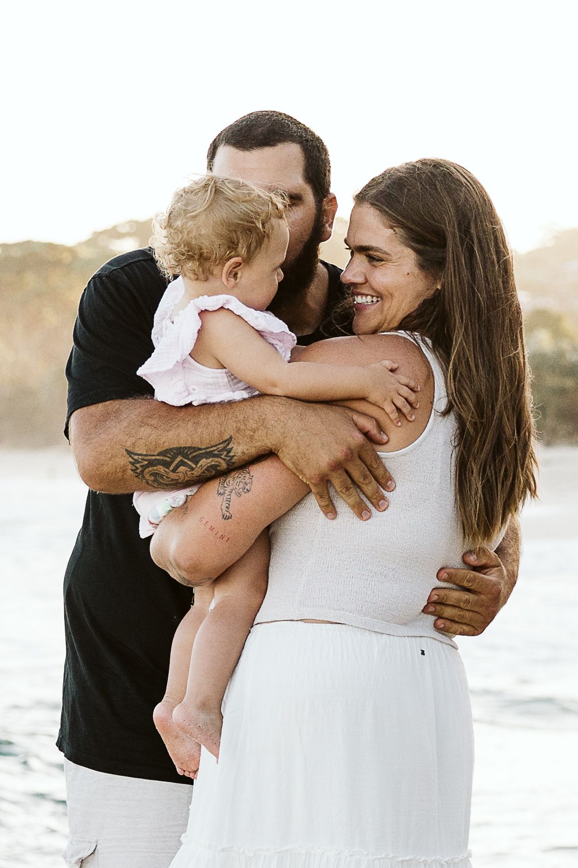 Happy family at beach, tattooed father kissing baby held by smiling brunette mother.