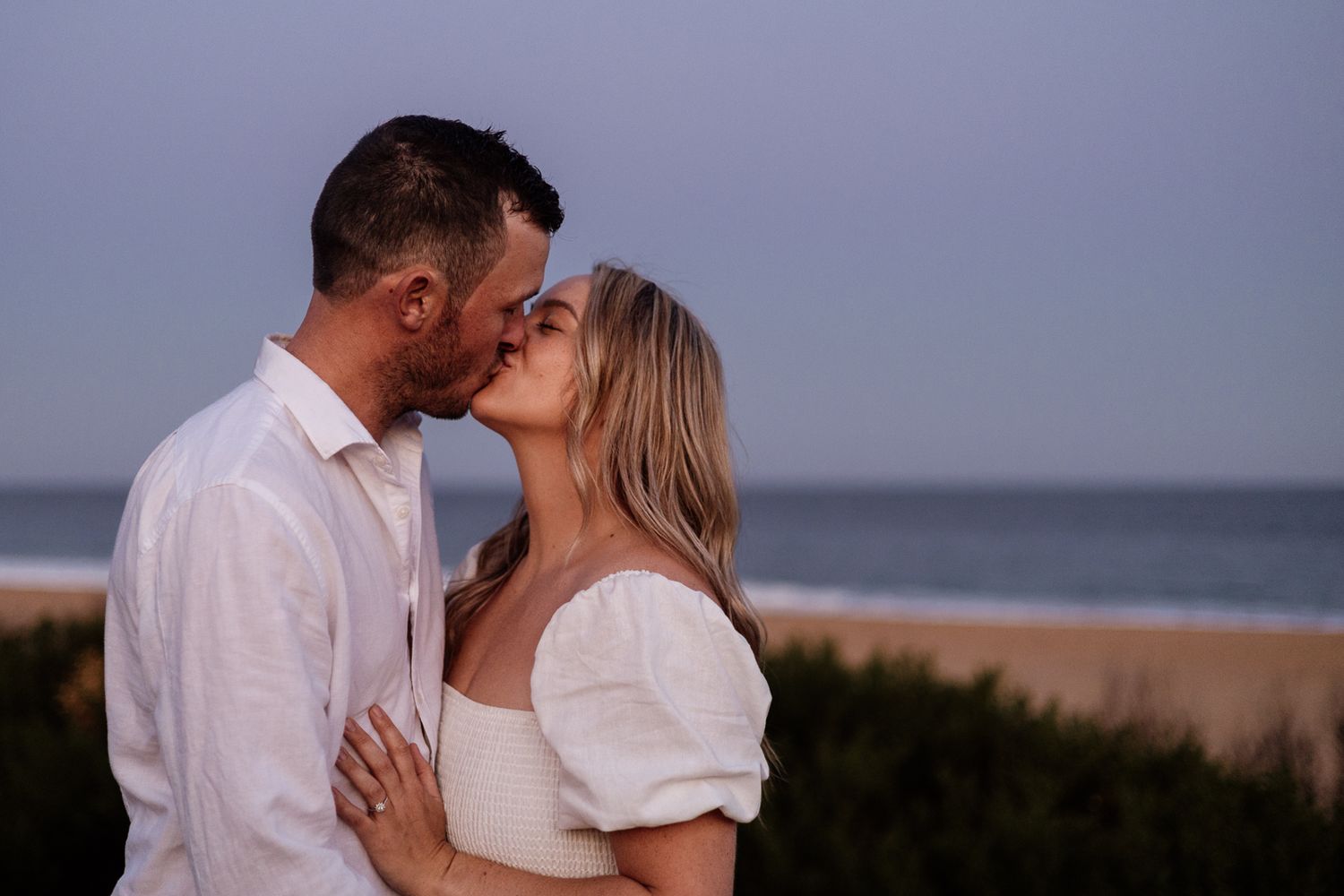 Couple sharing a romantic kiss with ocean beach backdrop at dusk.