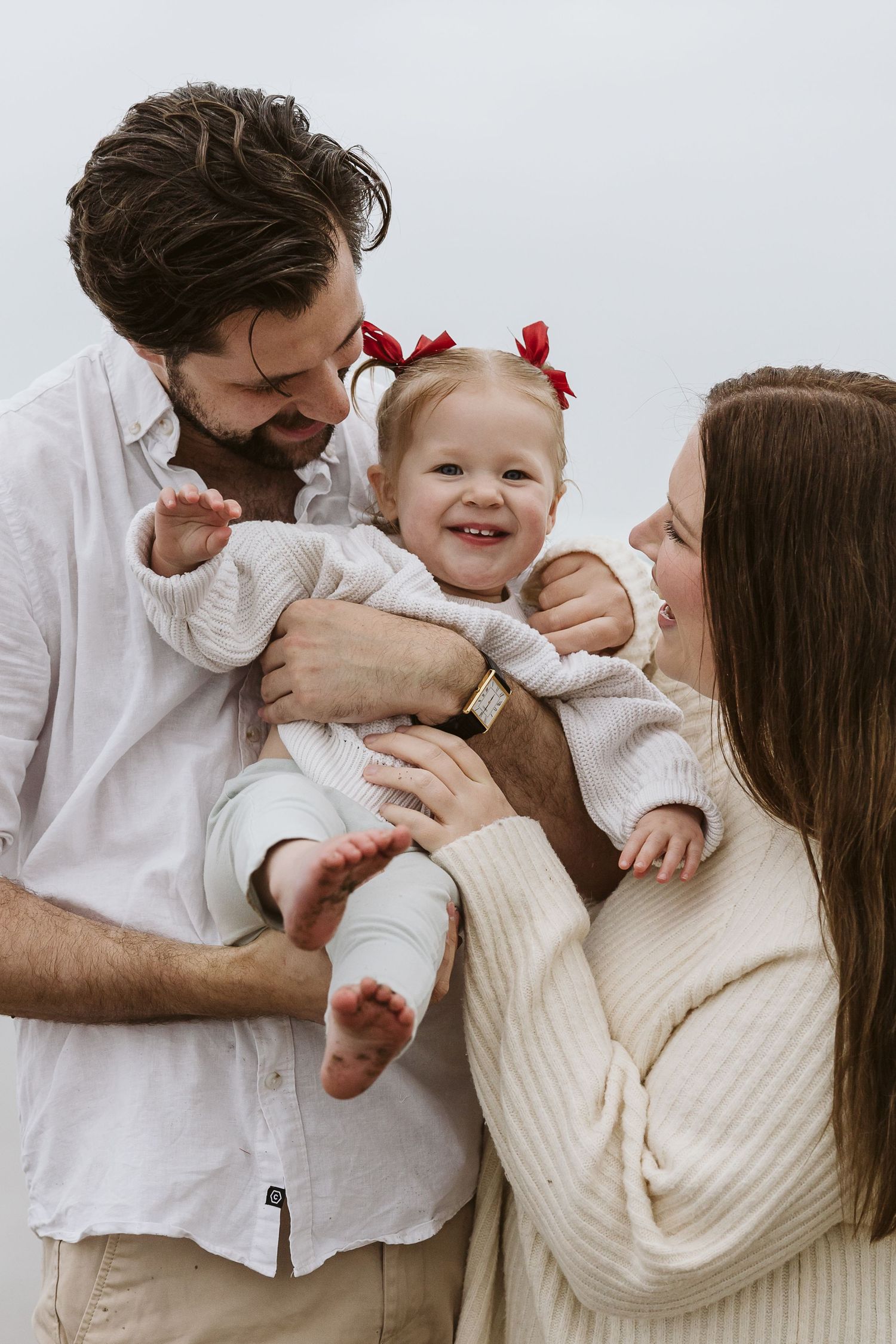 Happy family photo with parents holding smiling toddler girl with red bows outdoors.