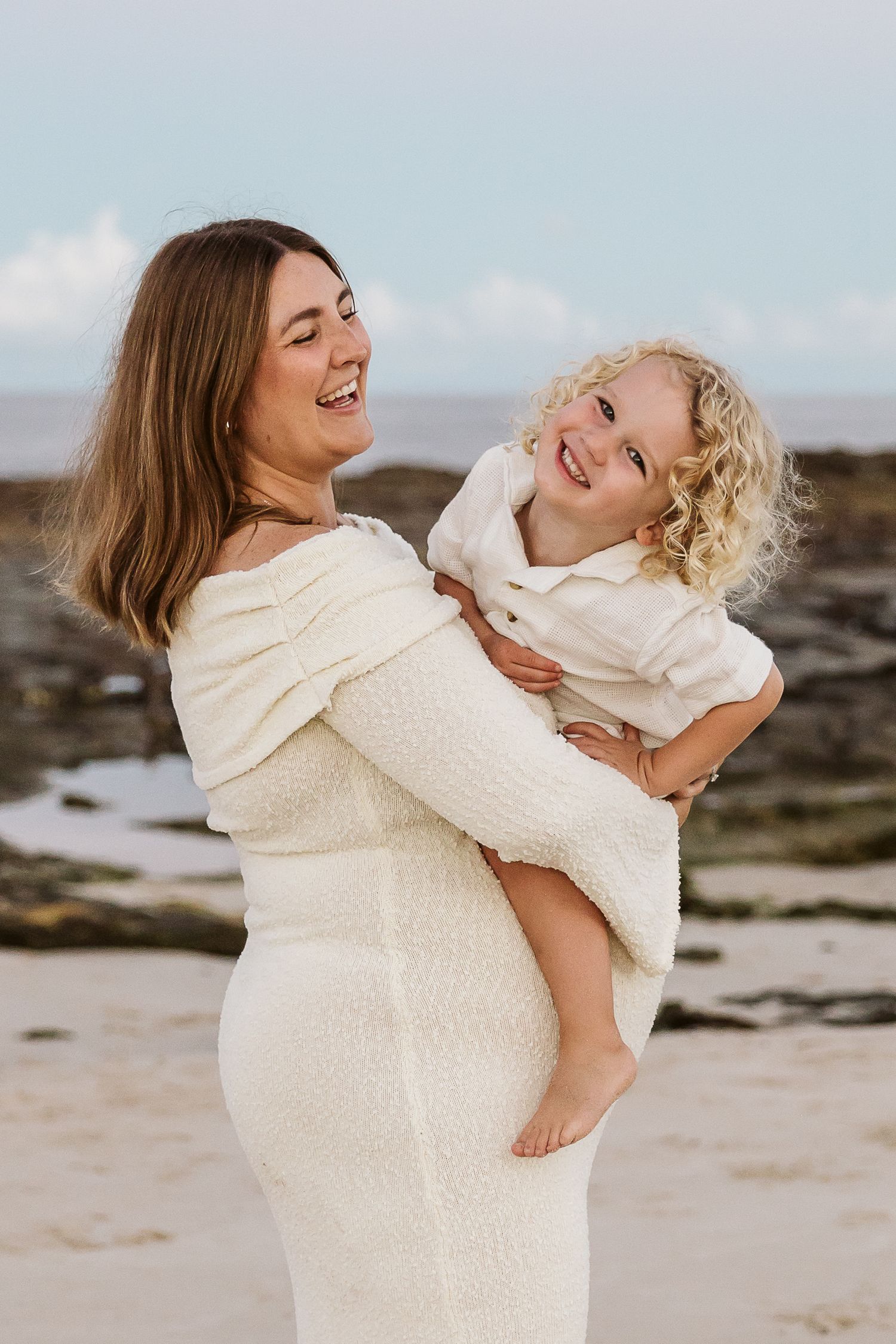 Laughing pregnant woman in cream knit dress holds smiling curly-haired toddler on rocky beach.