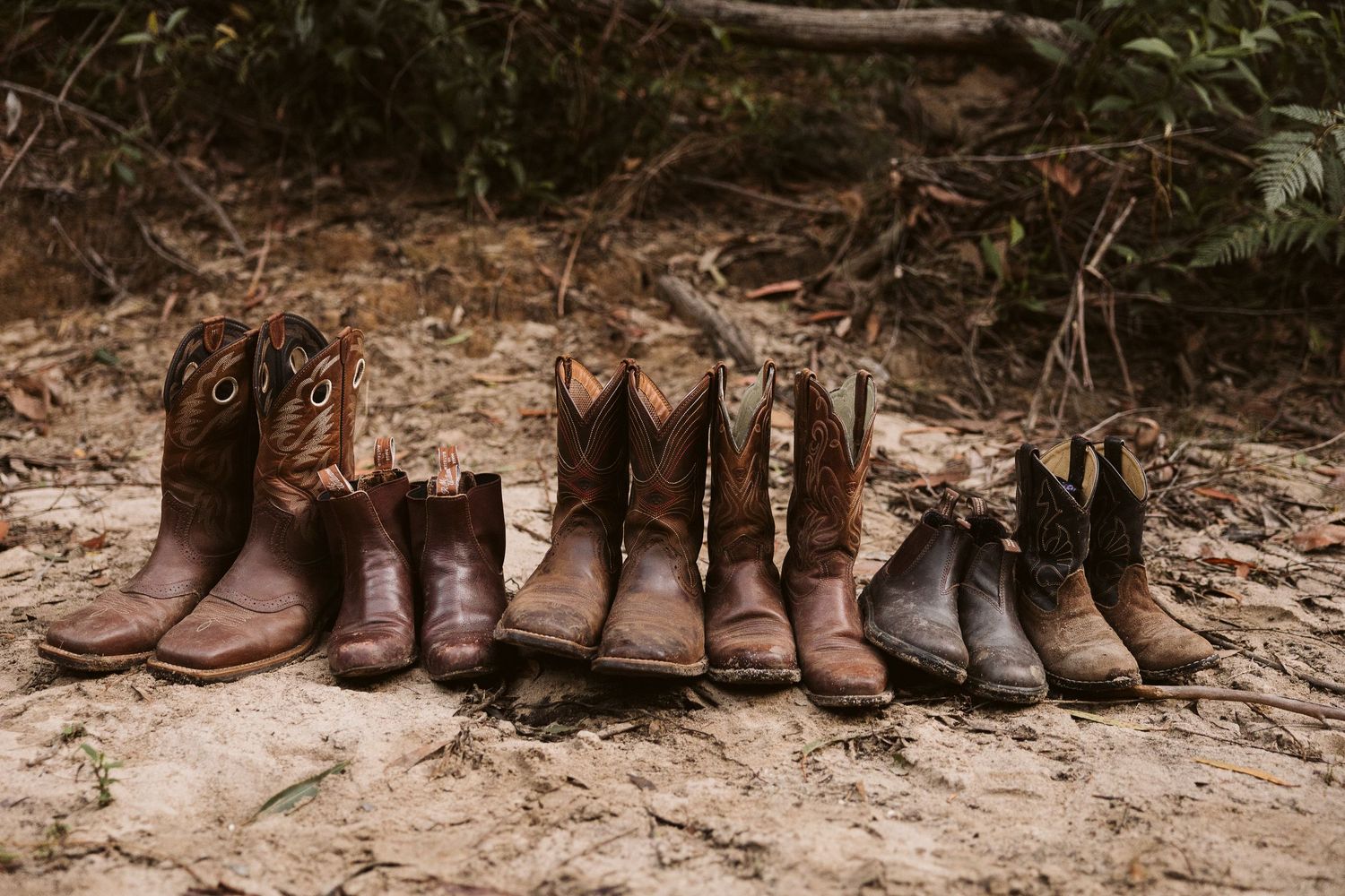 Five pairs of worn brown leather cowboy boots arranged in a row on sandy ground in the outdoors.