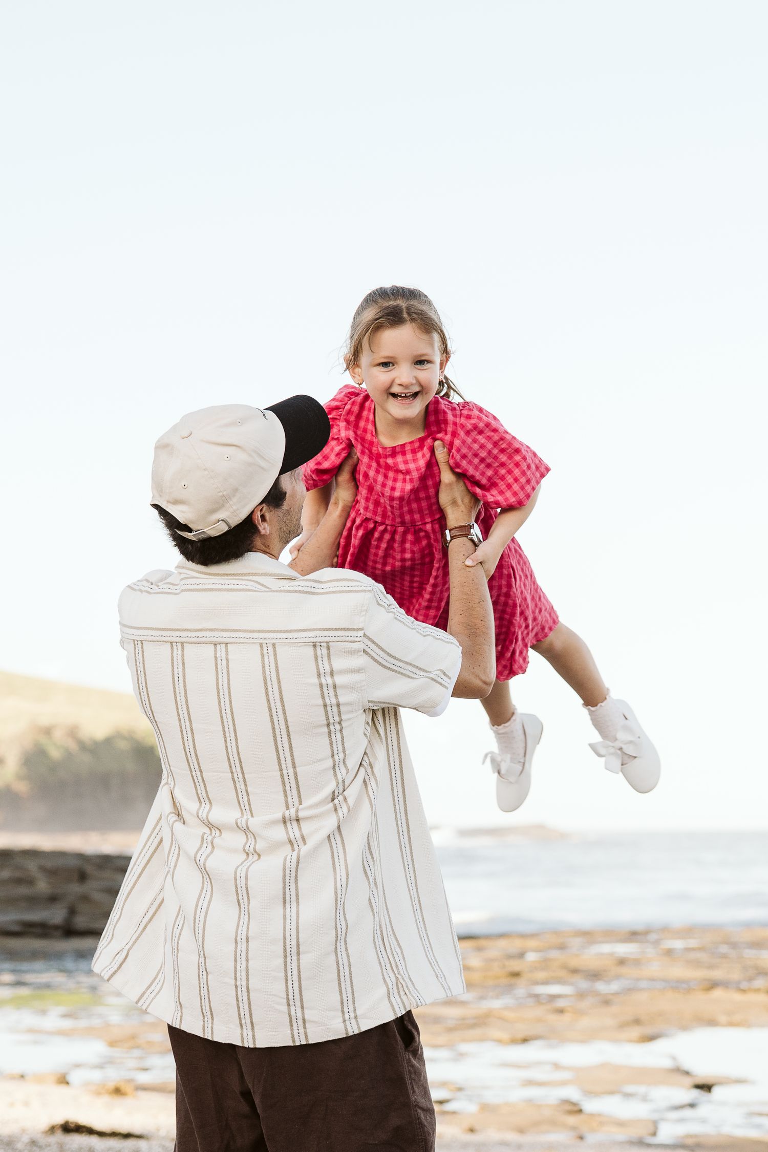 A man lifting a laughing girl in a red dress at the beach, sharing a joyful moment by the ocean.