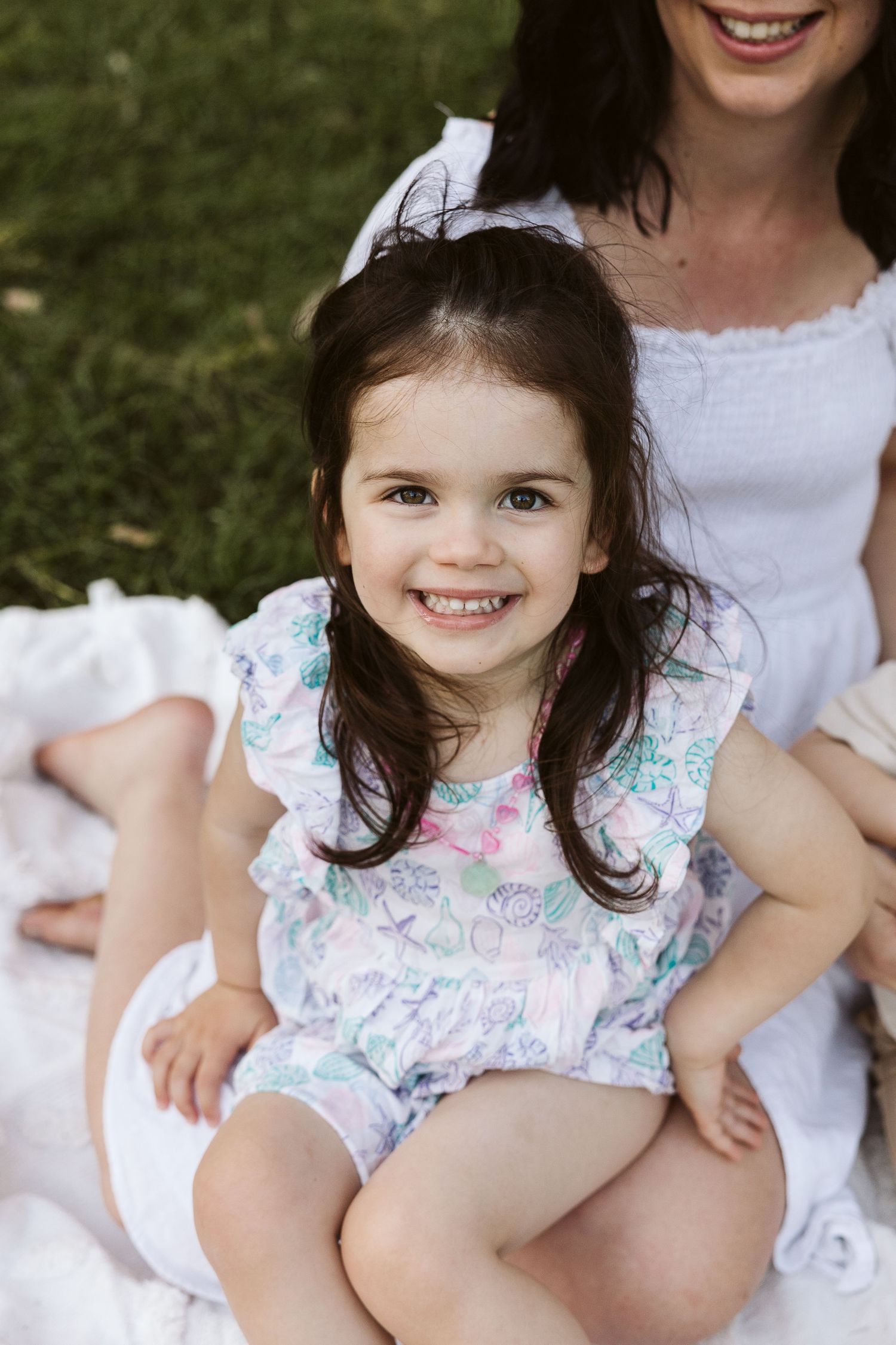 A smiling toddler girl with dark hair in a floral romper sits outdoors on a white blanket with her mother.