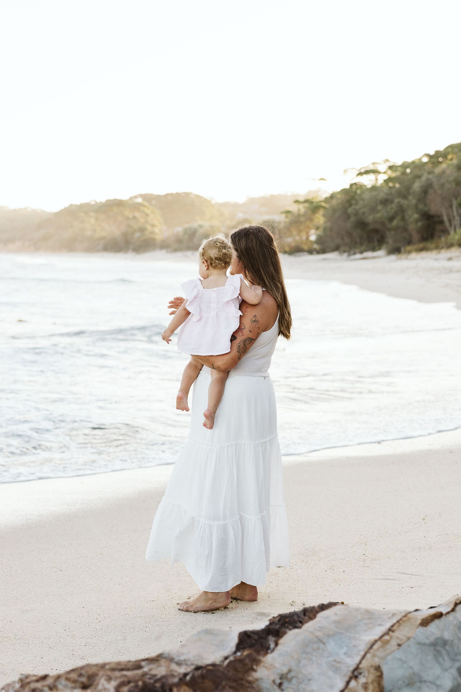 Mother in white dress holding toddler on sunny beach with ocean waves and green trees in background.