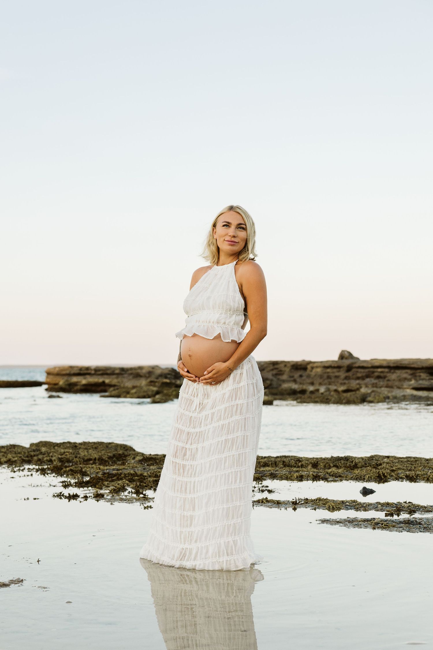 Blonde pregnant woman in white halter crop top and sheer maxi skirt standing on reflective beach at dusk.