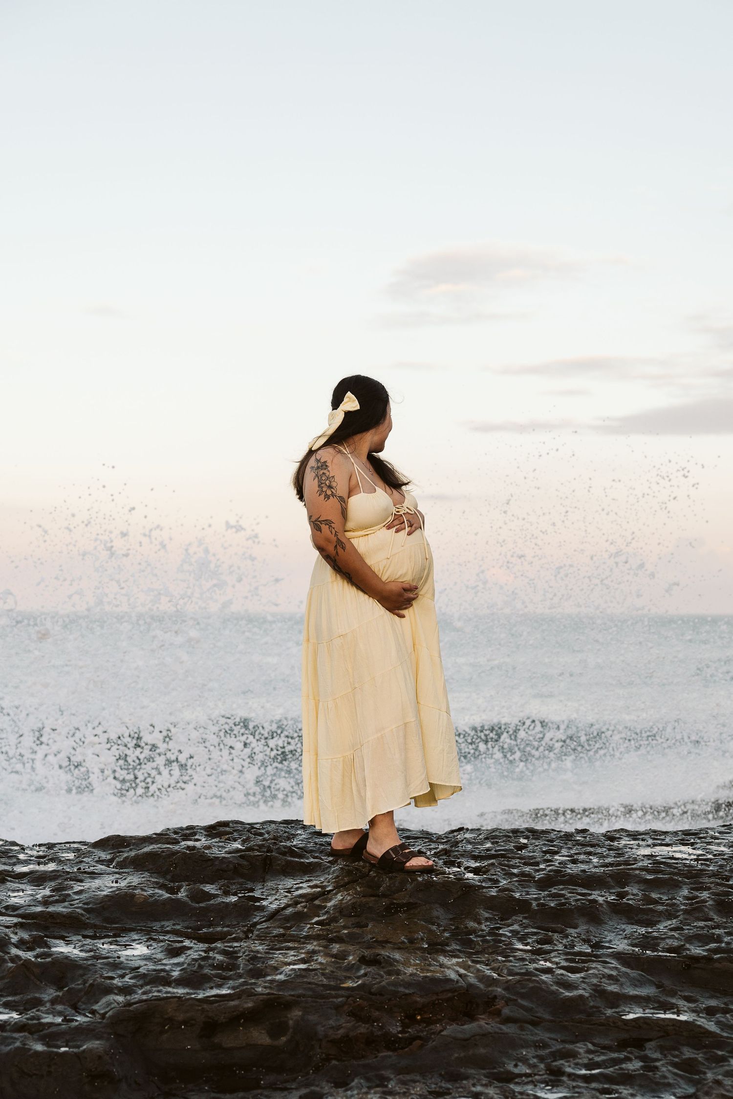 Pregnant woman in yellow dress standing on rocks by ocean waves at sunset.