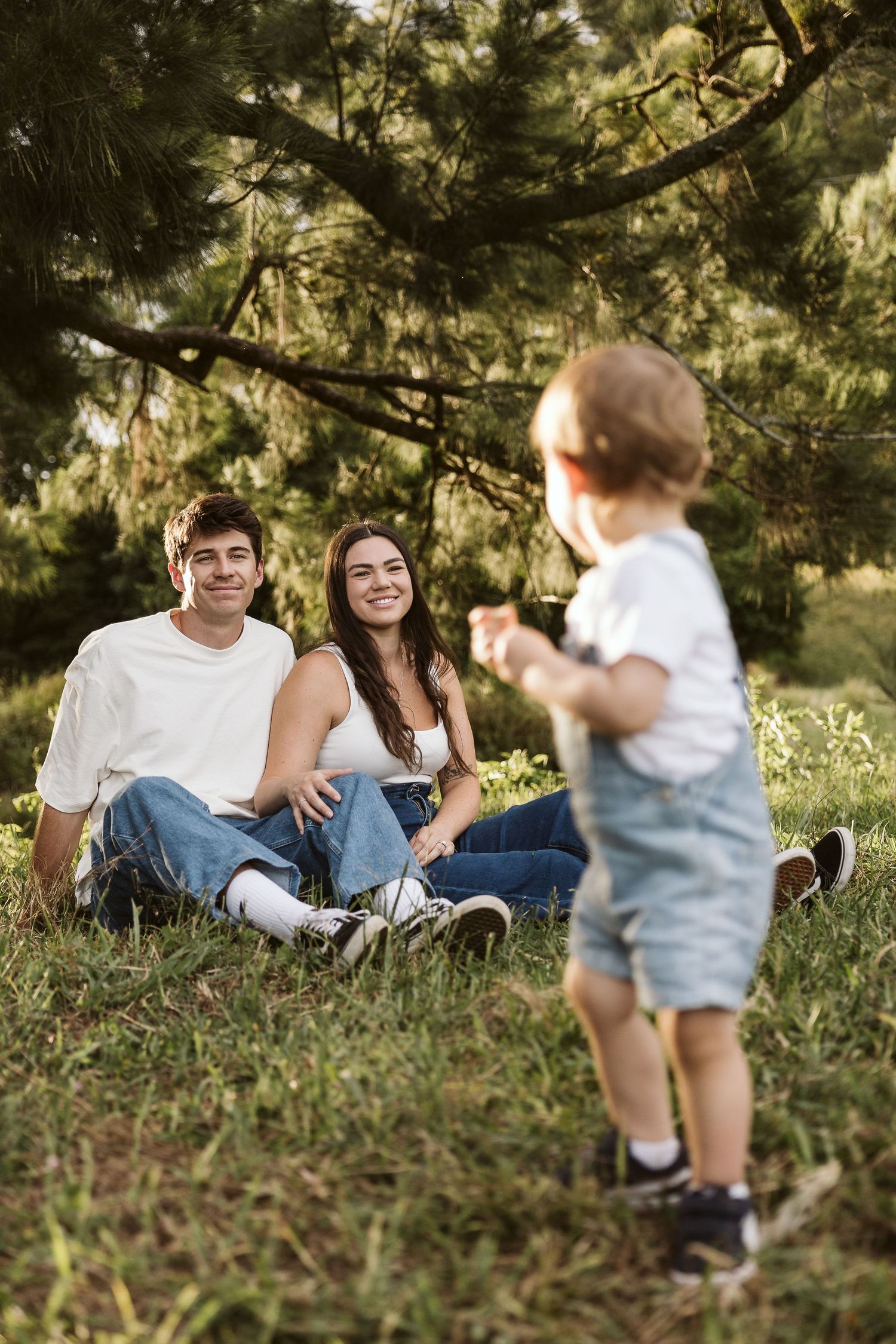 Young couple sitting on grass smiling at their toddler standing before them in a park.