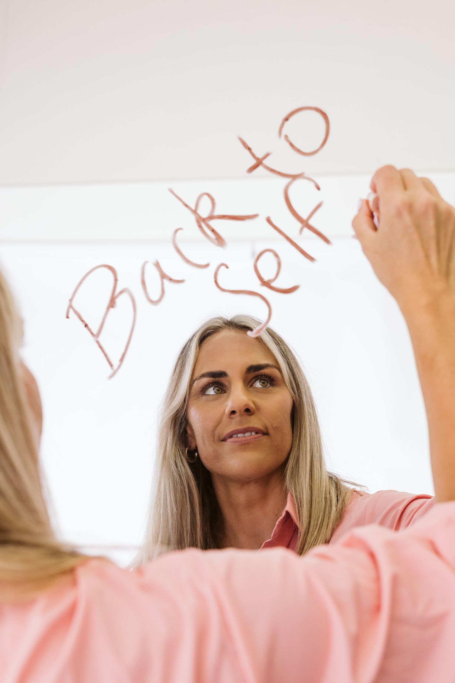 Woman in pink shirt writes Back to Self on mirror in lipstick, reflected image.