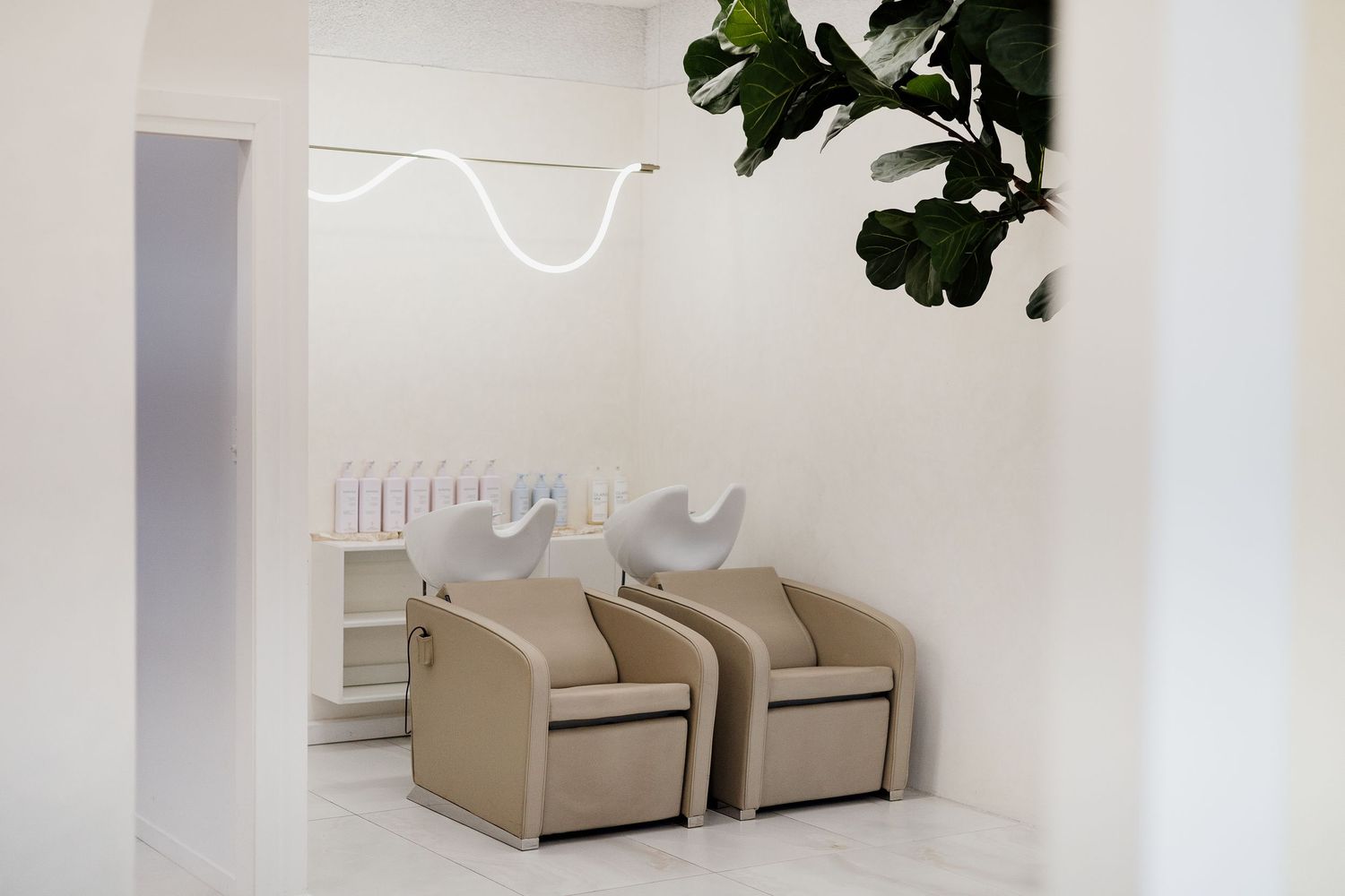 Modern hair salon wash stations with beige chairs, neon light fixture, and fiddle leaf fig plant.
