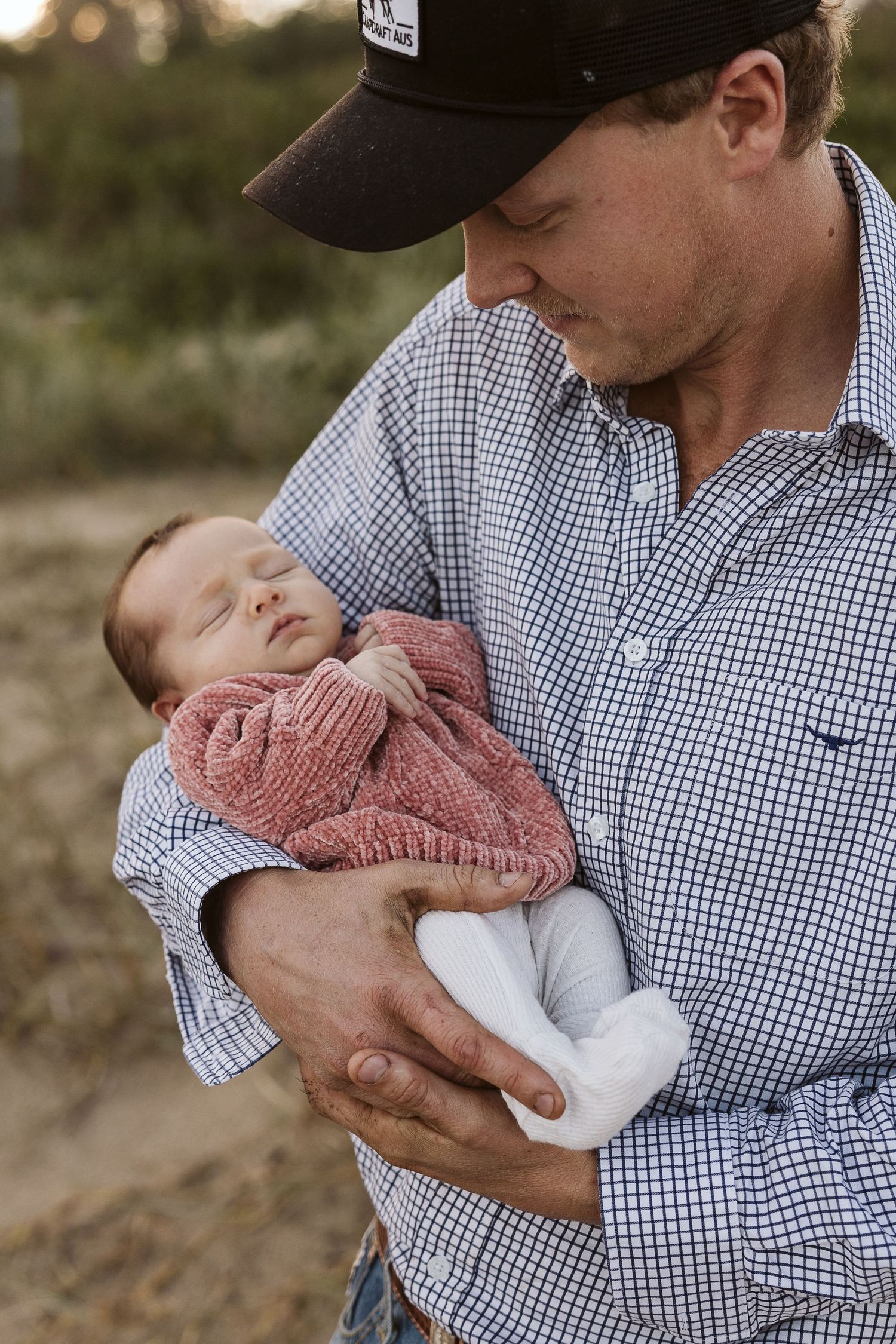 Father in plaid shirt and cap tenderly cradling sleeping newborn in pink knit outfit.
