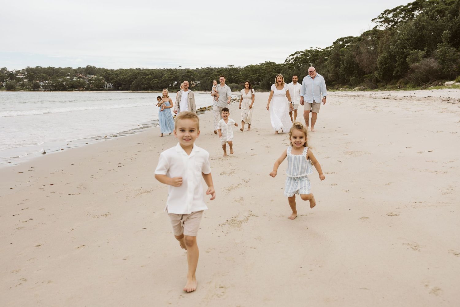 Children run joyfully on a sandy beach while a large family walks together in matching white and blue outfits.