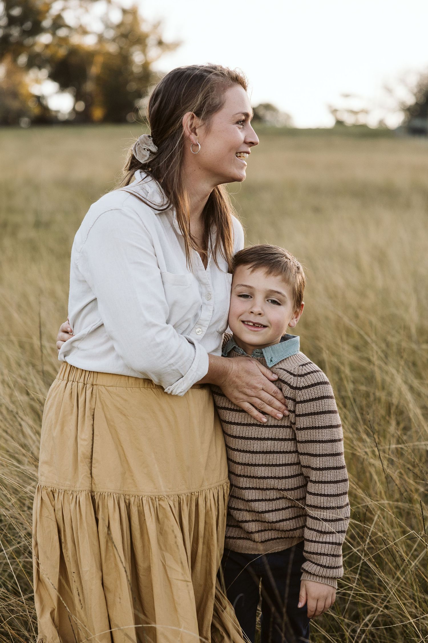 A smiling woman in a white blouse and golden skirt embraces a young boy in a striped sweater in a golden grass field.