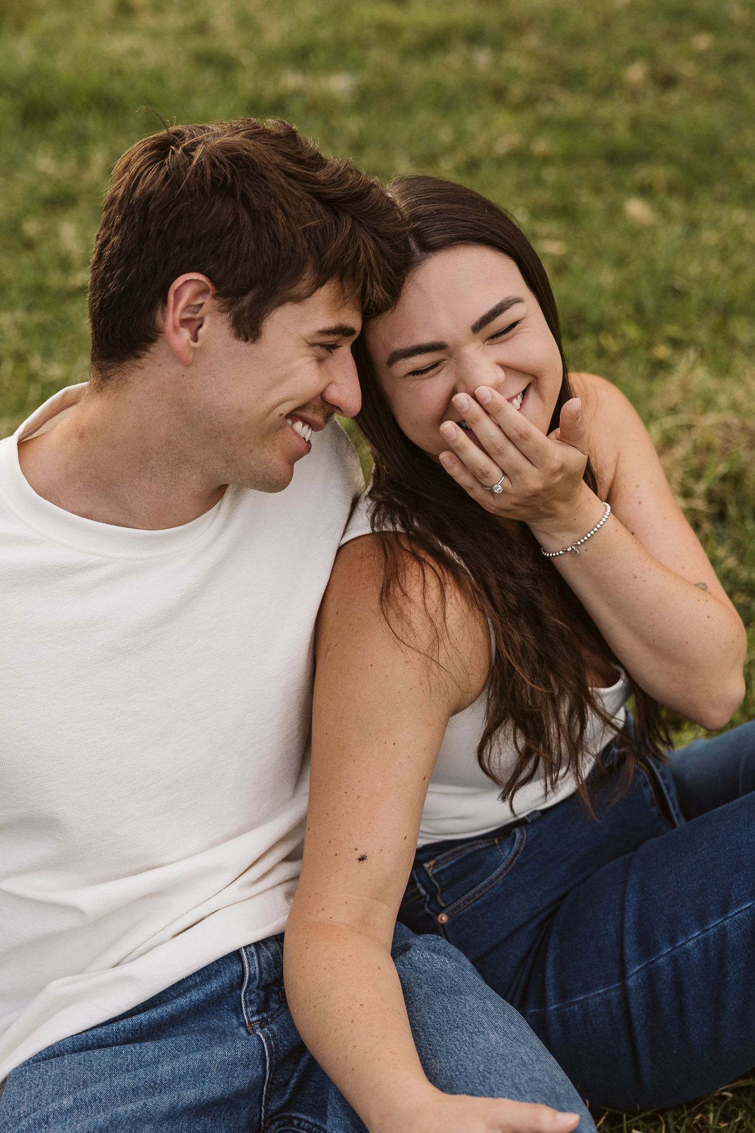 Couple laughing together while sitting on grass, woman covering her smile, both in white tops and blue jeans outdoors.