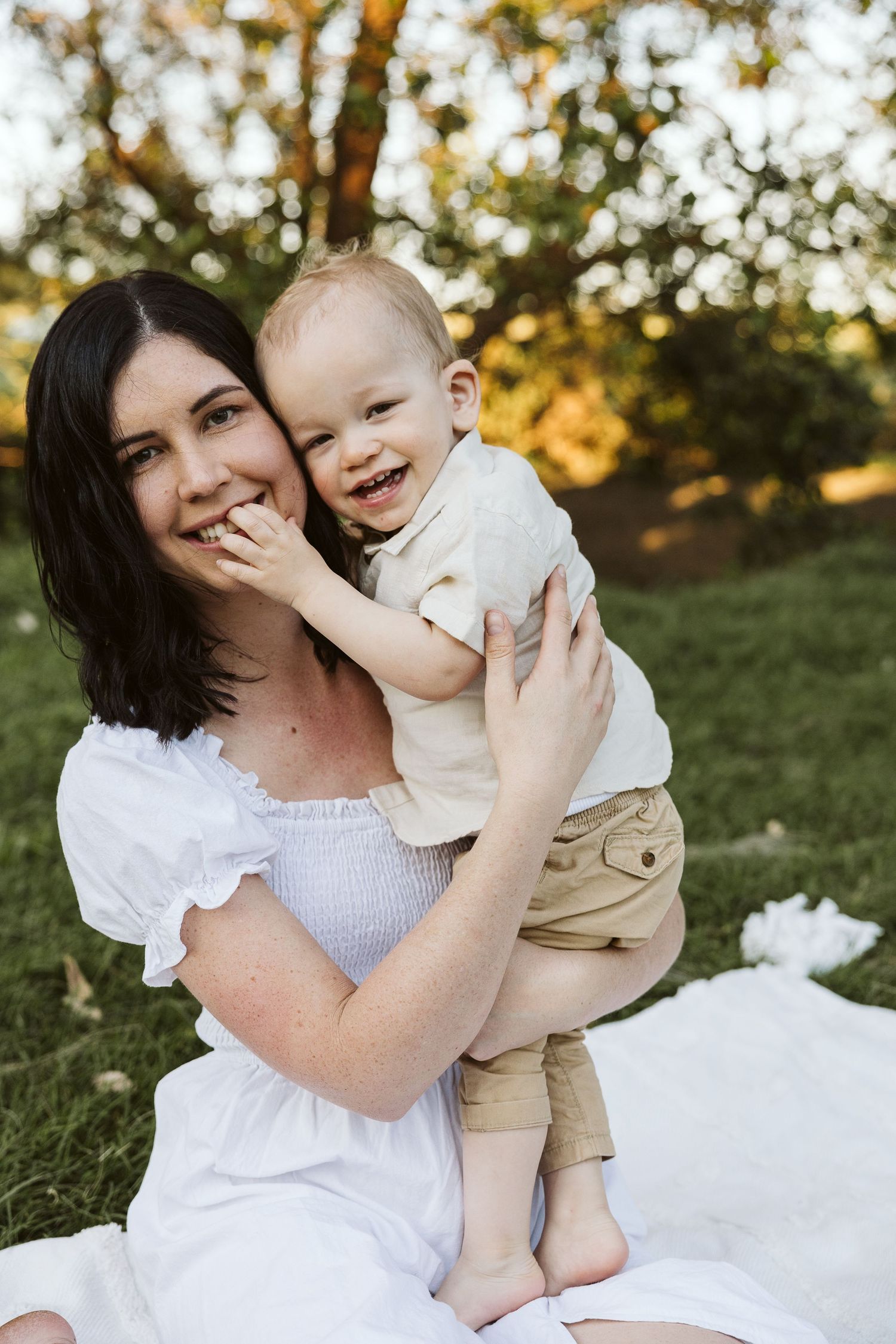 A smiling mother in a white dress holds her laughing baby boy outdoors on a sunny day.