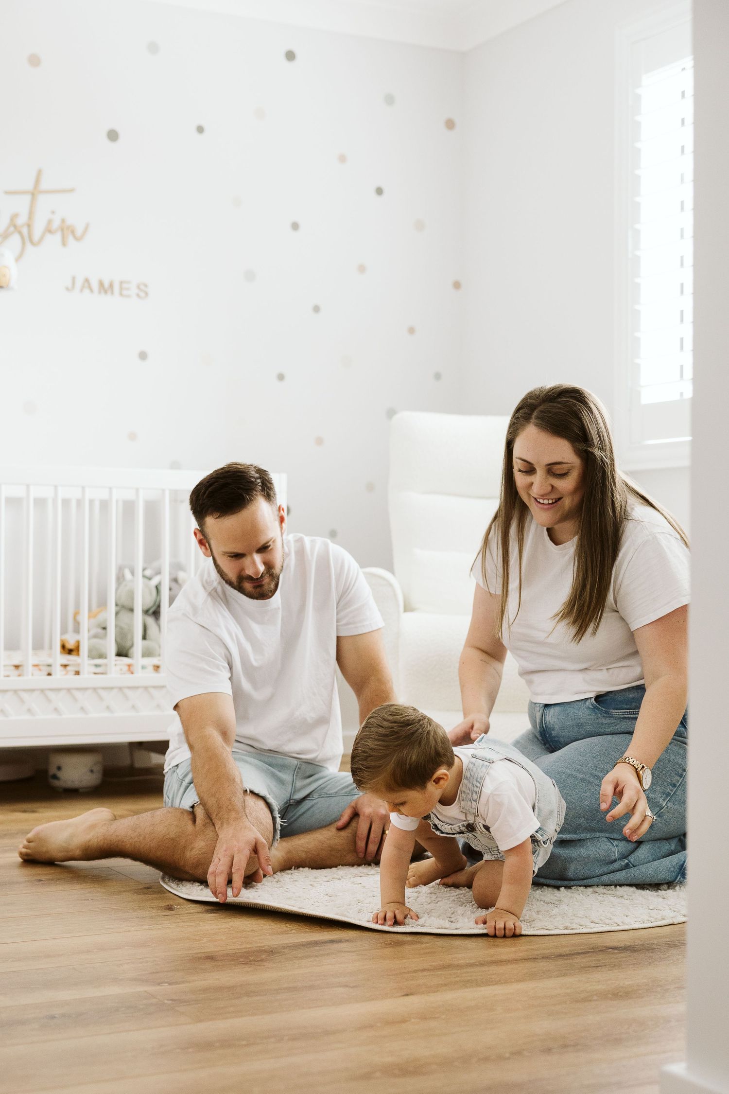 Parents in white tees and jeans sit on nursery floor with crawling baby, polka dot walls and white crib behind them.