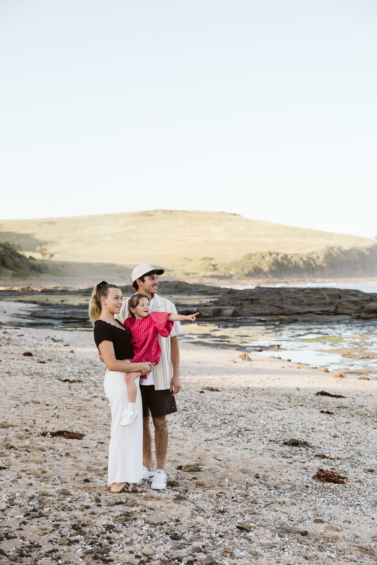 A young family with a toddler in red stands together on a rocky beach with rolling hills behind them.