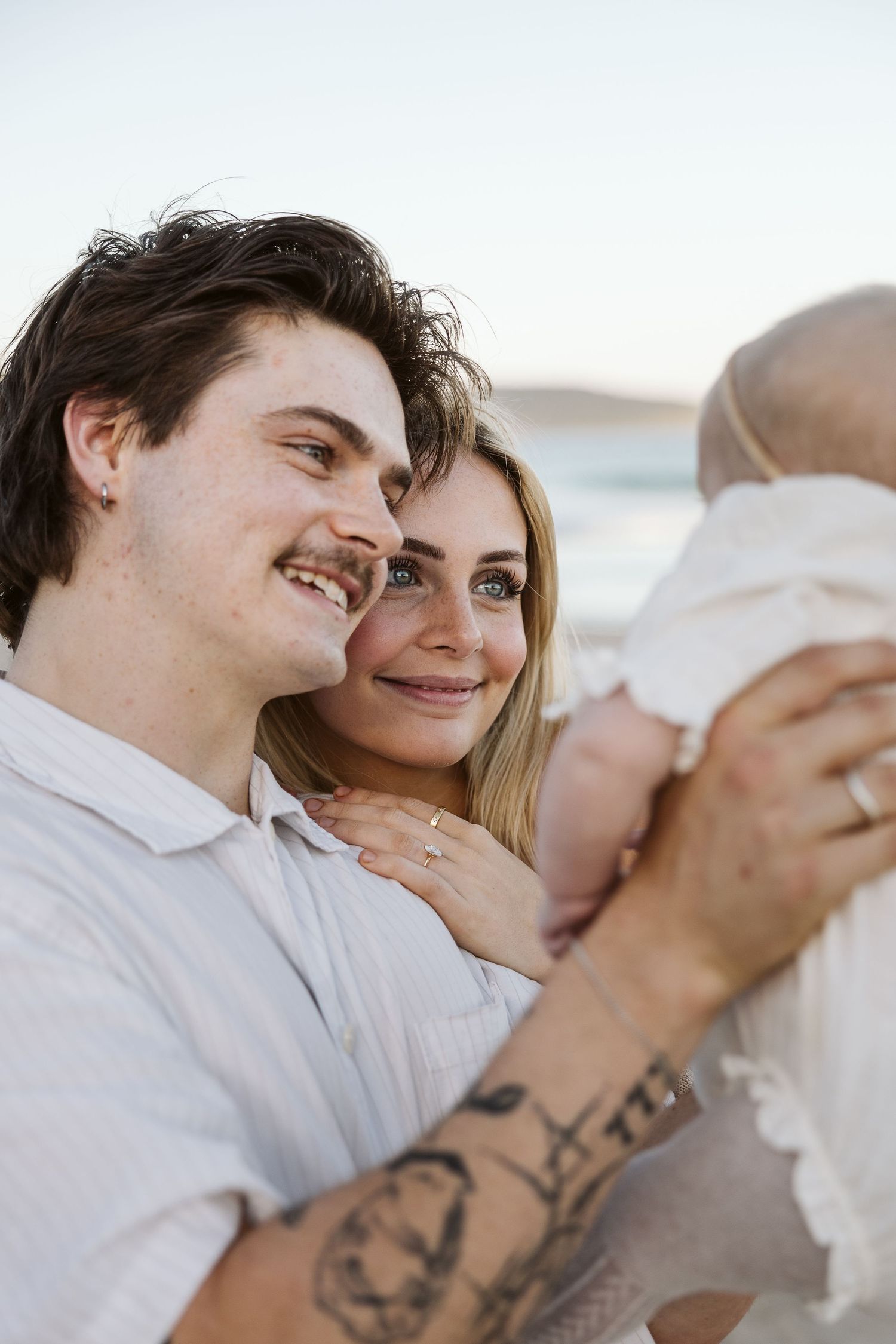 Young couple smiling and holding their baby at the beach during golden hour.