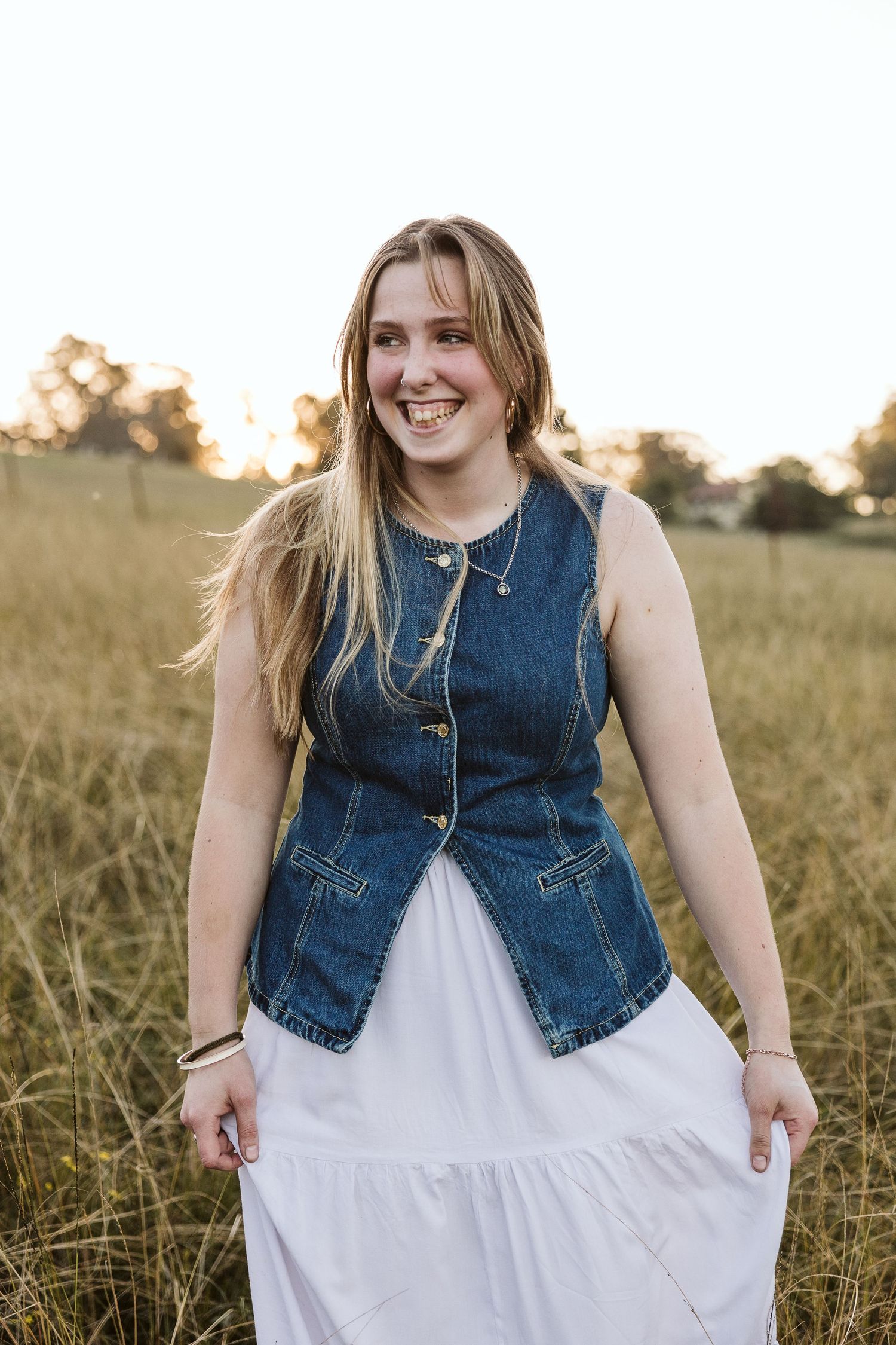 Smiling young blonde woman in a denim vest and white dress standing in a sunlit golden grass field.