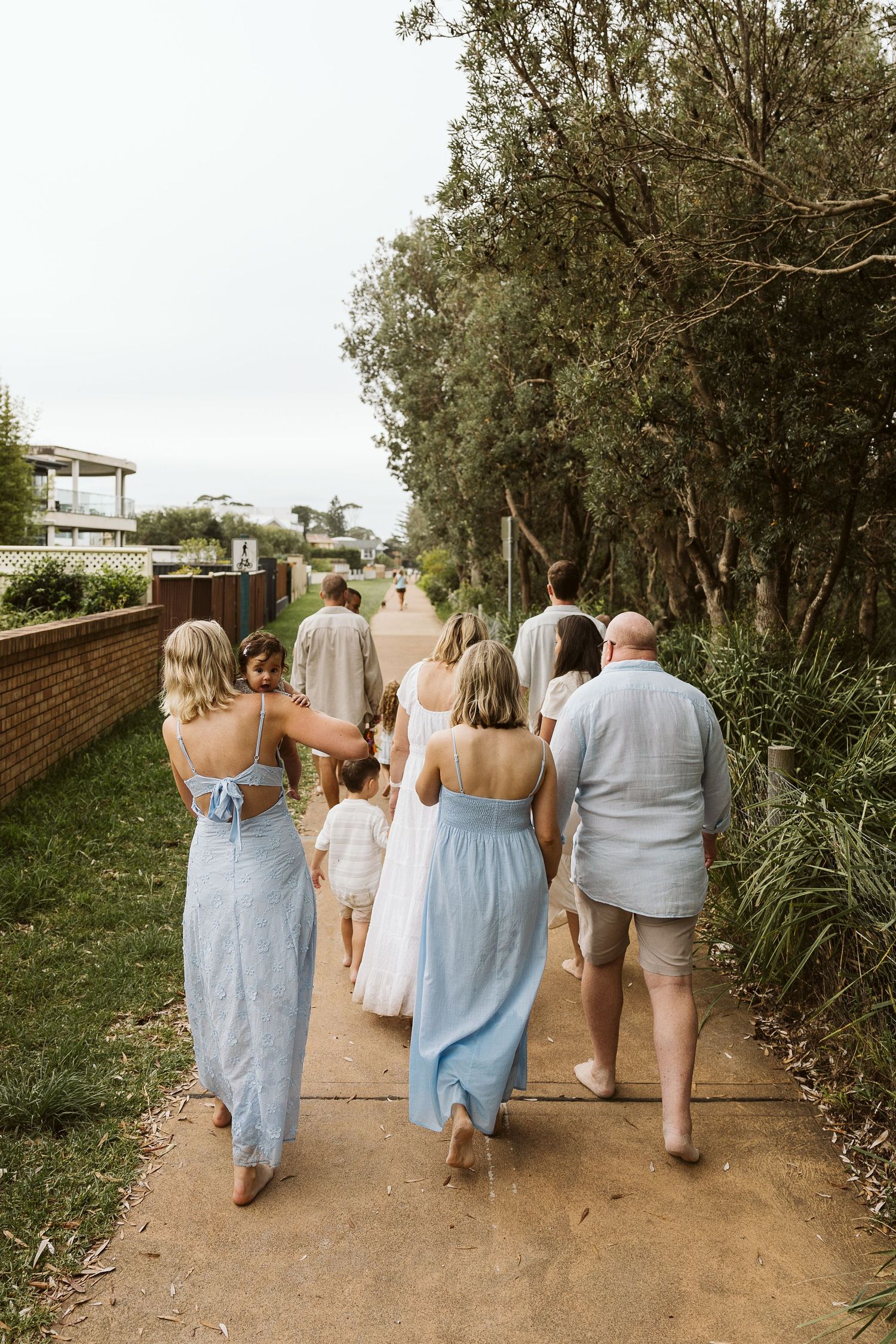 Group of family members walking along a sunny coastal path lined with green trees and a brick wall.