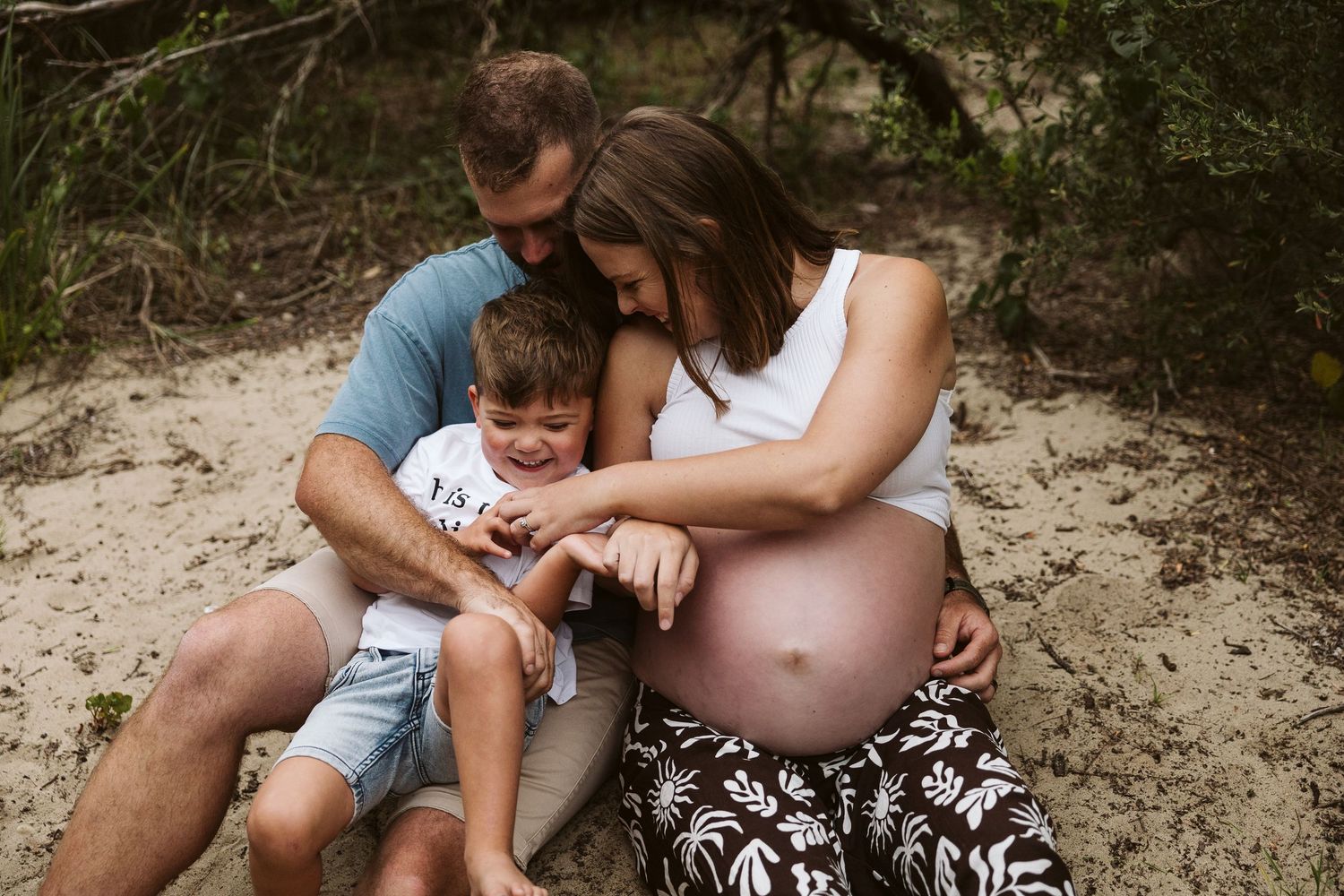 Expecting couple sitting with young son in sandy wooded beach area.