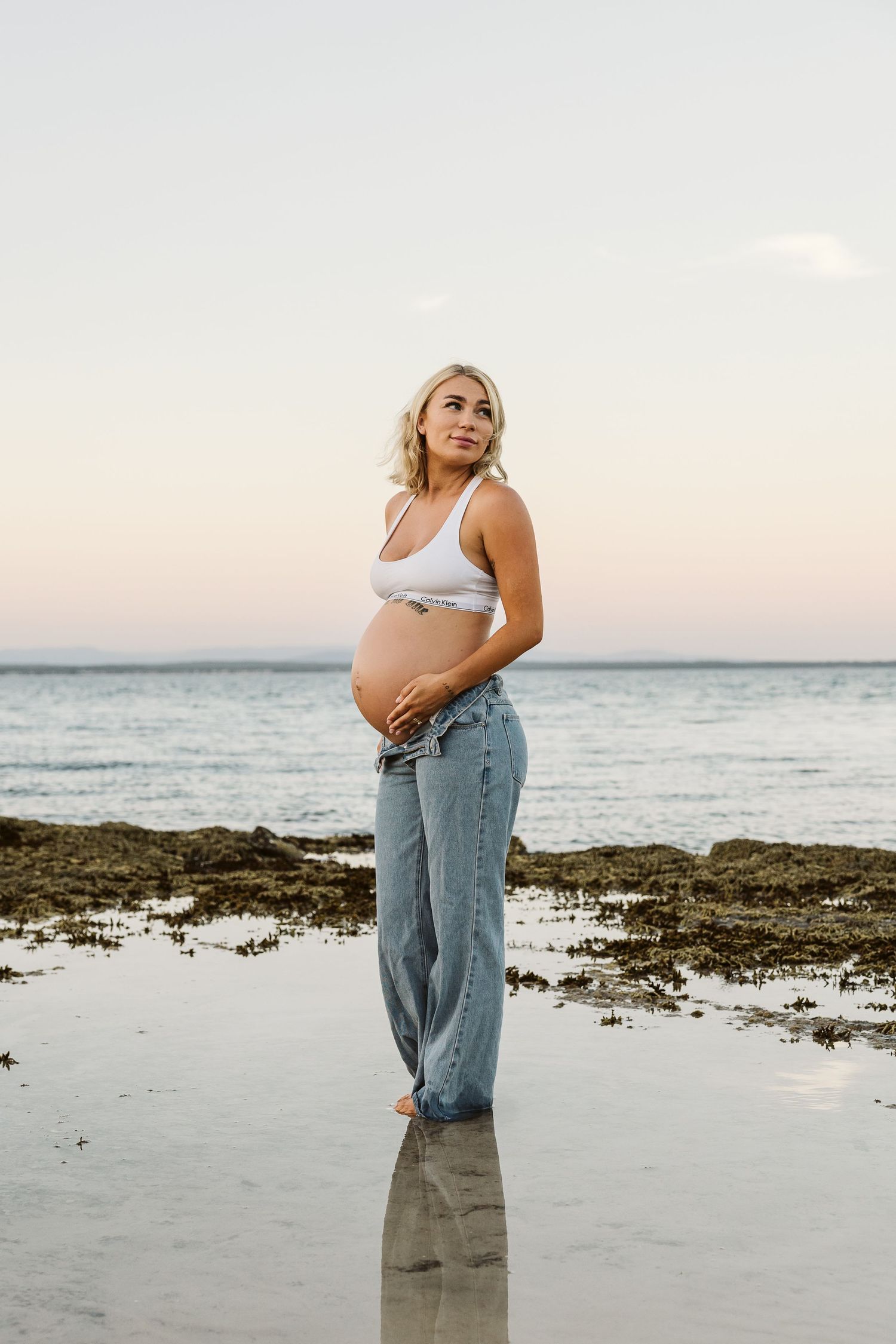 Blonde pregnant woman in white sports bra and wide-leg jeans standing on reflective rocky beach at sunset.