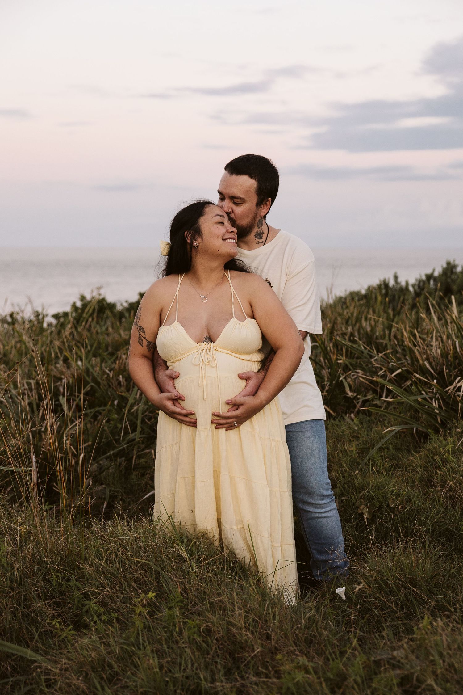 Man embracing pregnant woman from behind in grassy coastal field at sunset.