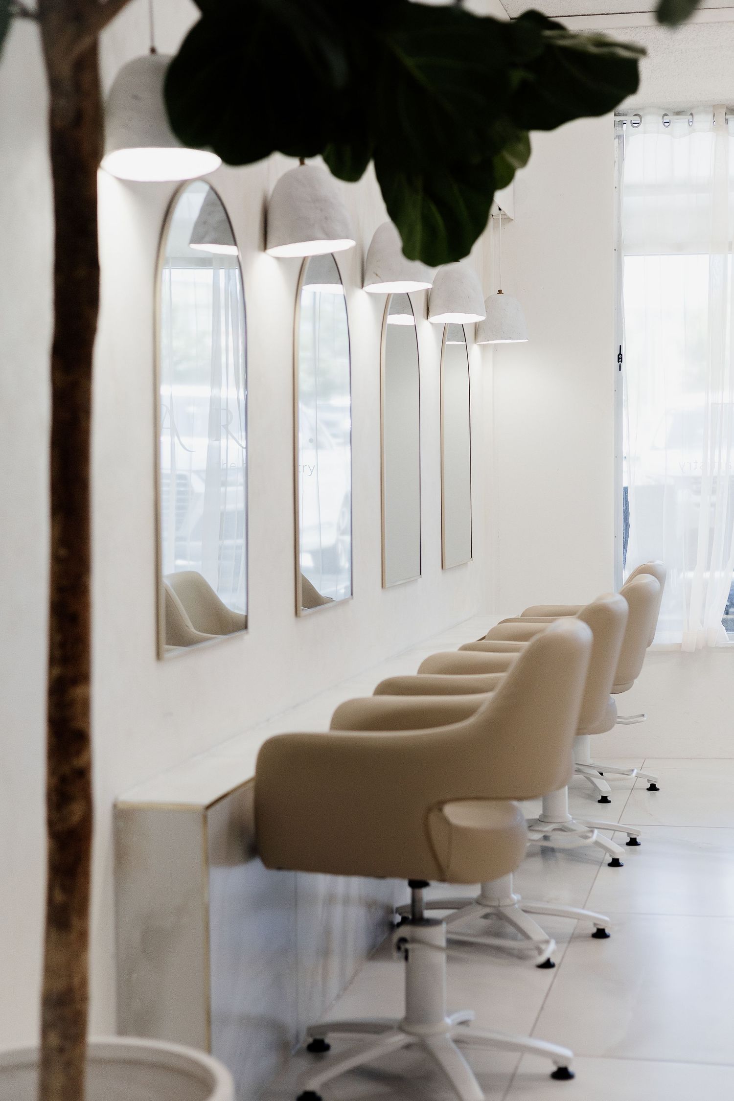 Modern hair salon interior with beige styling chairs and arched mirrors along a white wall.