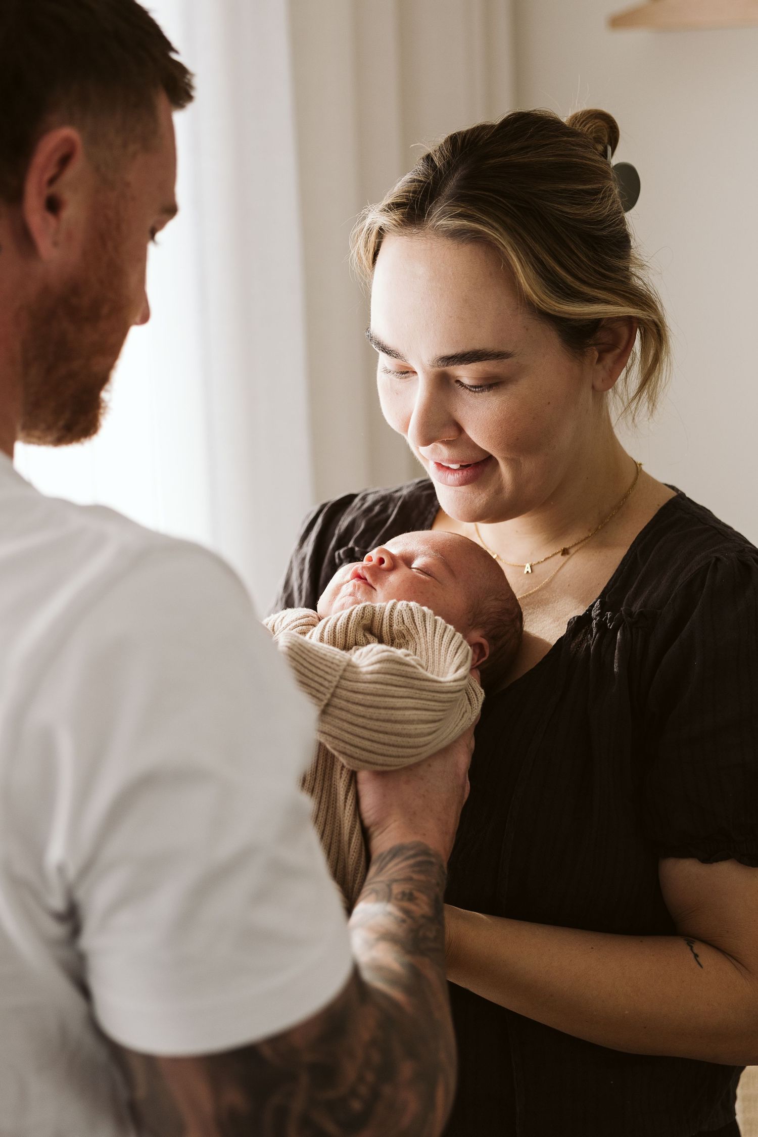 New parents smiling and holding their newborn baby wrapped in a knit hat indoors.