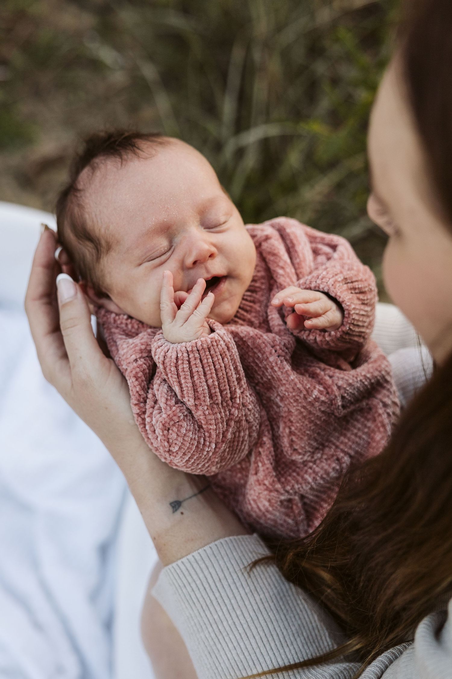 Mother holding smiling newborn baby dressed in pink knit sweater outdoors.