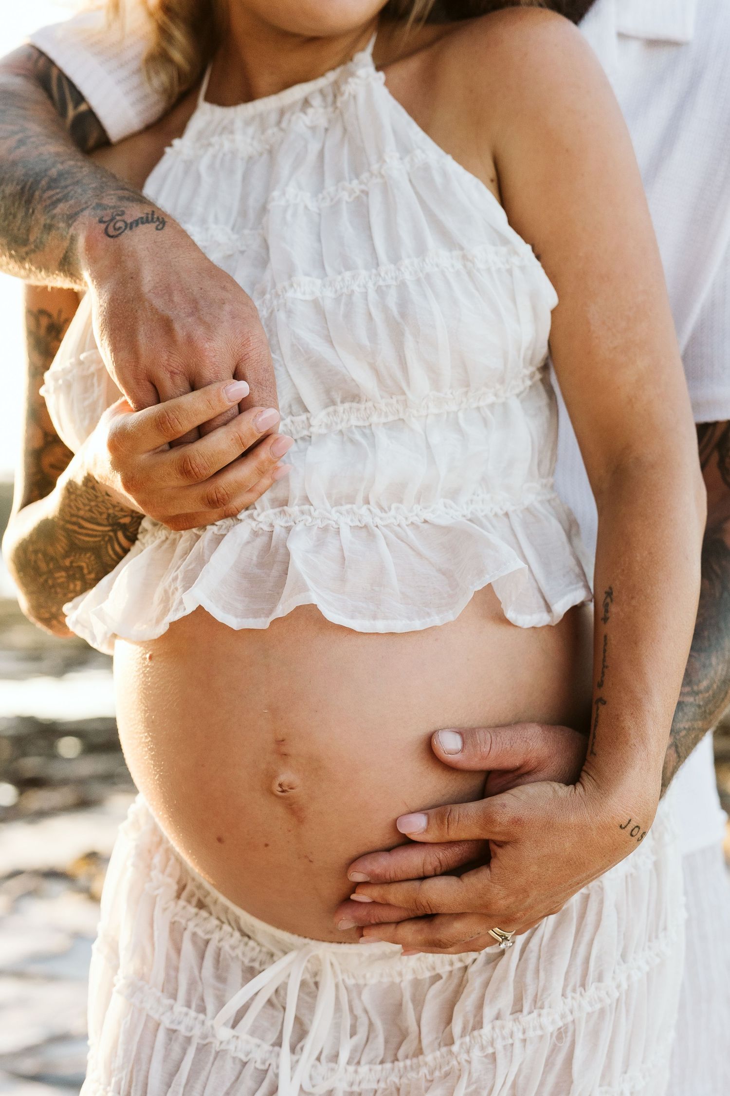 Close-up of tattooed man's hands embracing pregnant woman's bare bump in white ruffled halter top outdoors.