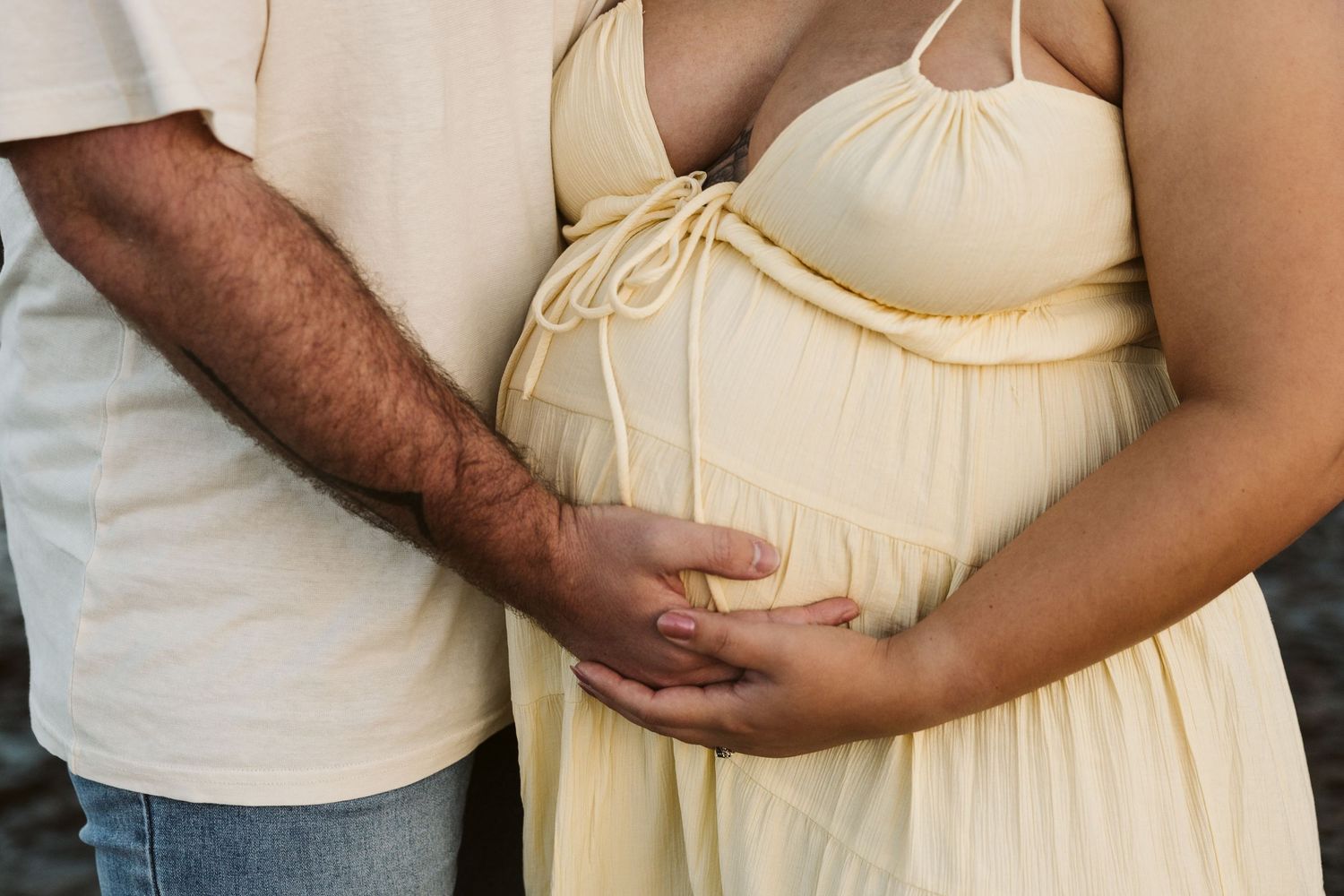 Close-up of couple holding pregnant belly together, woman in yellow maternity dress.