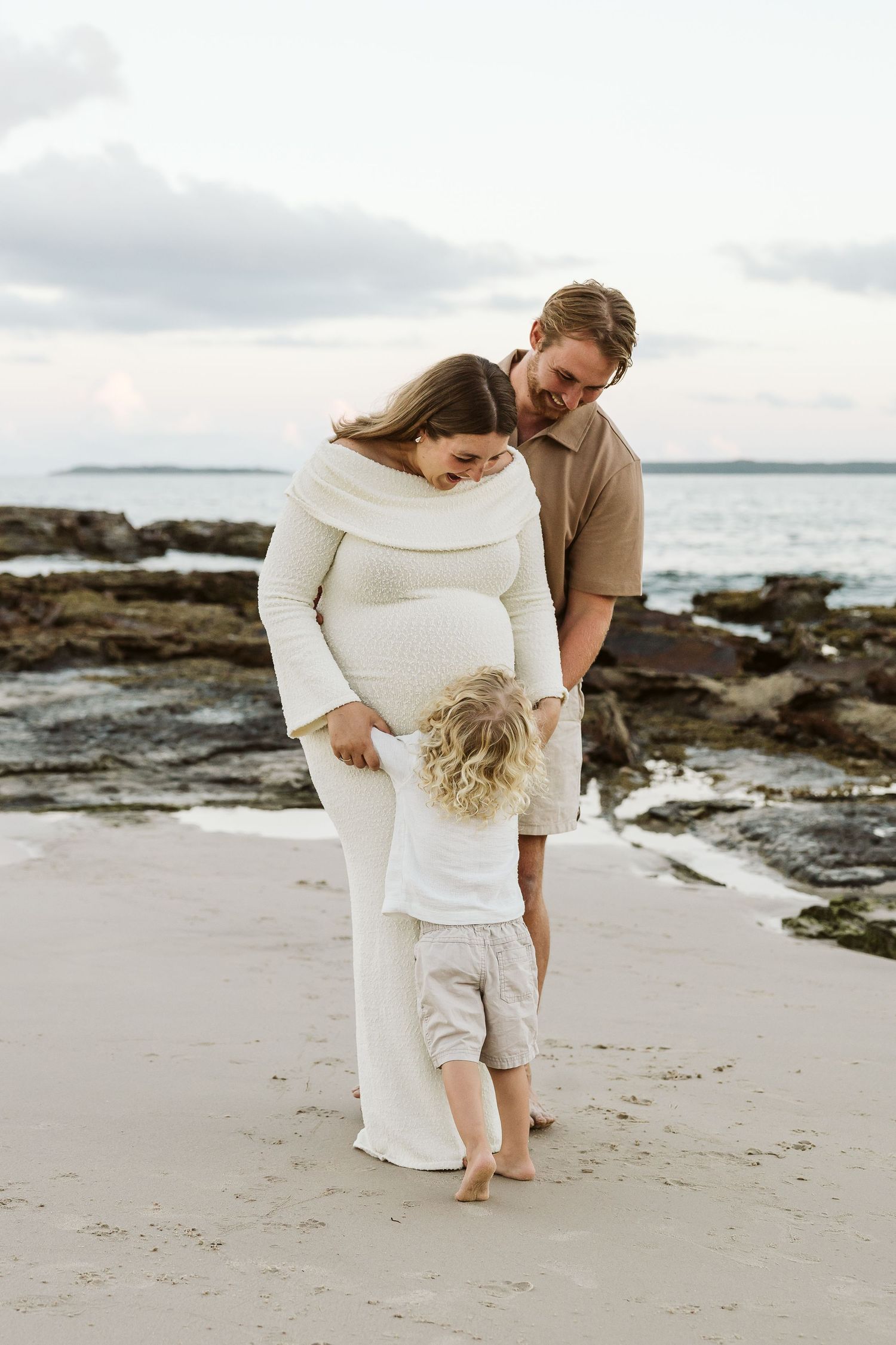 Pregnant woman in cream knit dress with partner and curly-haired toddler on sandy beach at dusk.