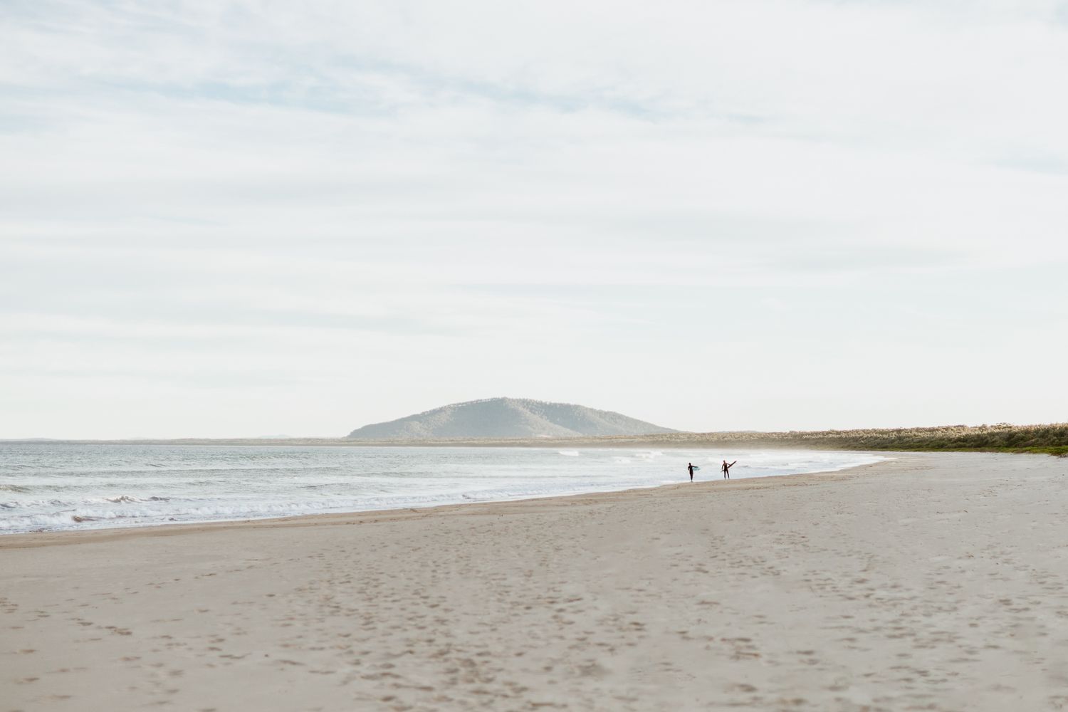 Wide open sandy beach with calm ocean waves and distant hill under overcast sky.