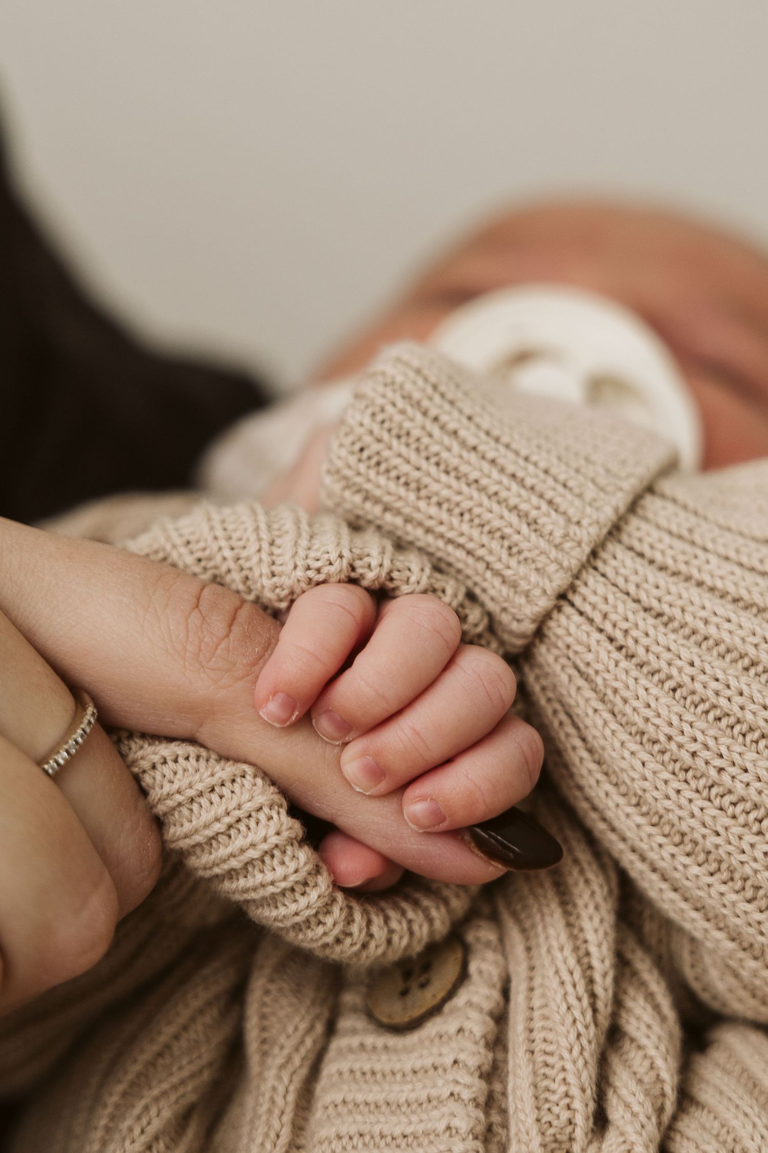 Adult hand with ring gently holding tiny newborn baby fingers in a knit sweater.