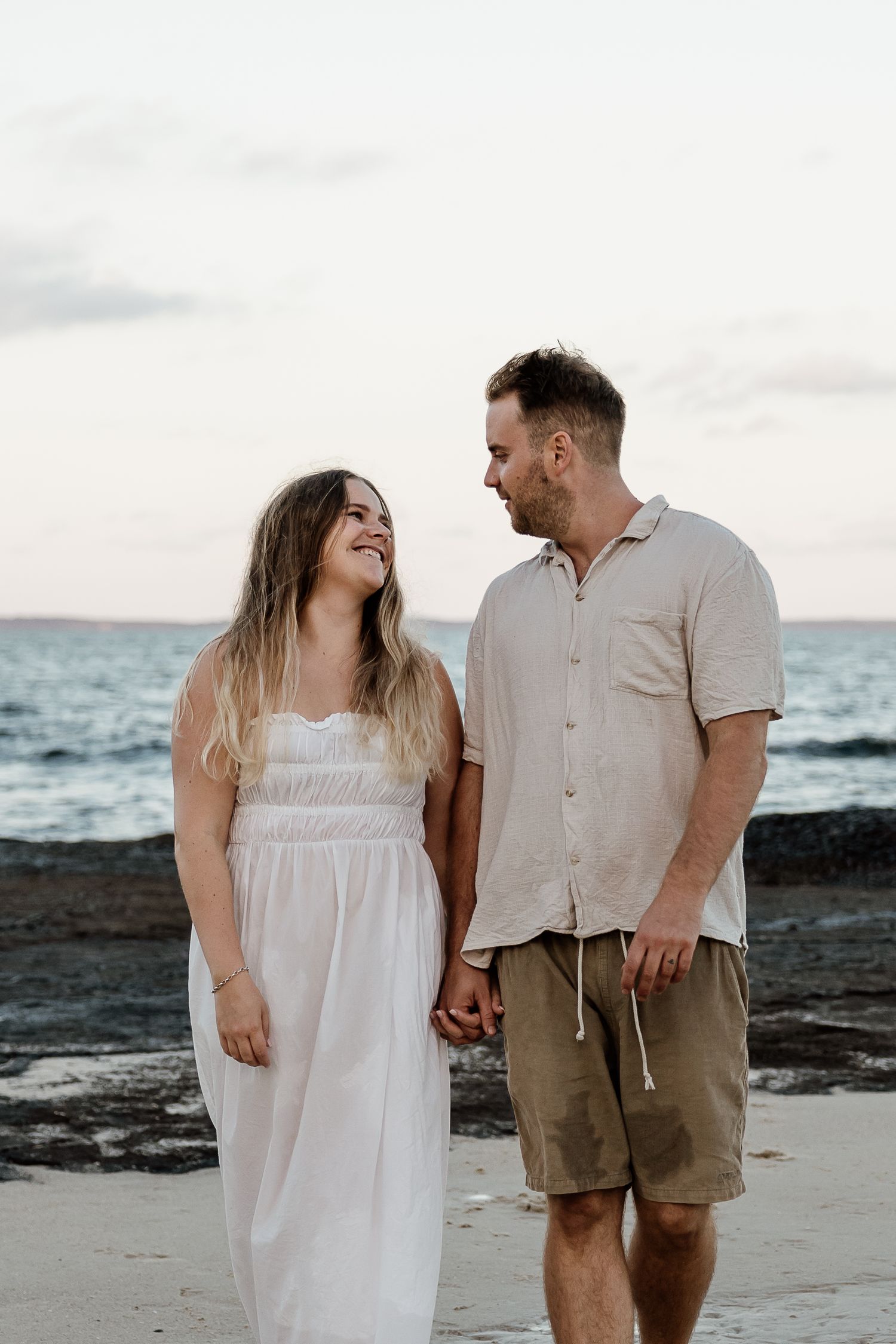 Couple holding hands and smiling at each other on a rocky beach at sunset, woman in white dress, man in beige shirt.