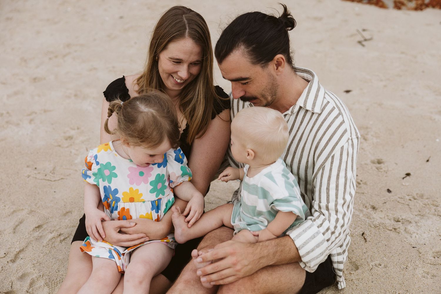 Young family of four sitting together on sandy beach laughing with two toddlers.