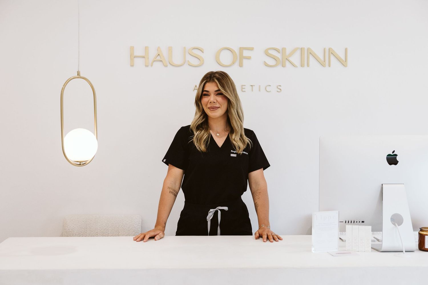 Female aesthetician in black scrubs standing confidently behind white reception desk at Haus of Skinn Aesthetics salon.