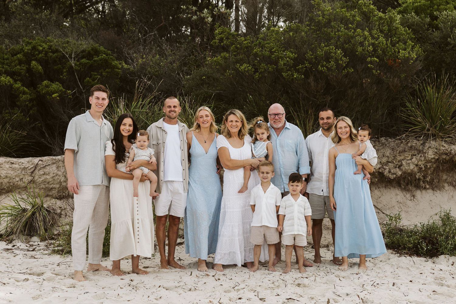 Large family posing together on a sandy beach in coordinated light blue and white outfits with lush green trees behind.