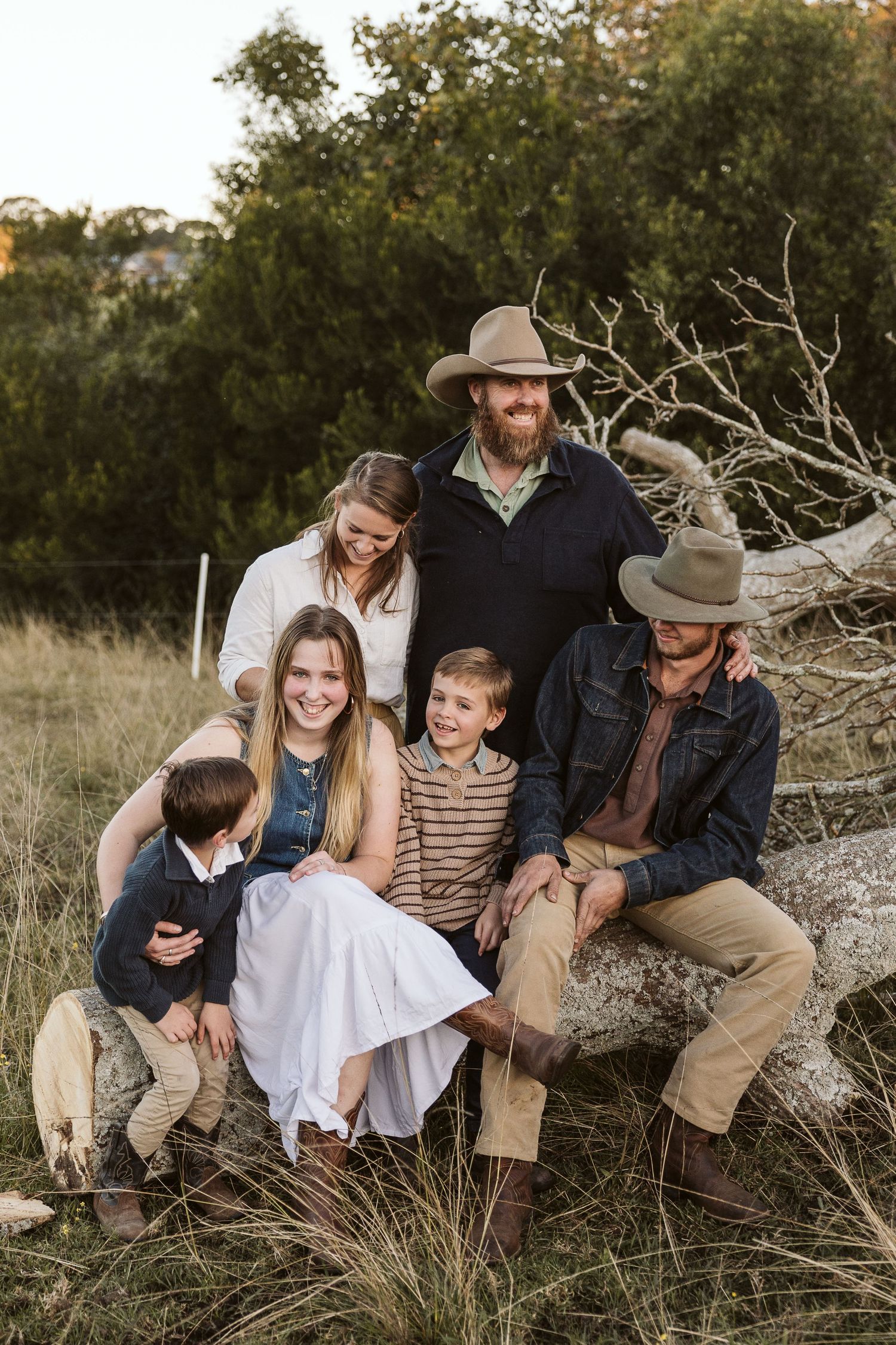 A happy family of five poses outdoors on a fallen log in a grassy field, wearing country-style clothing and cowboy hats.