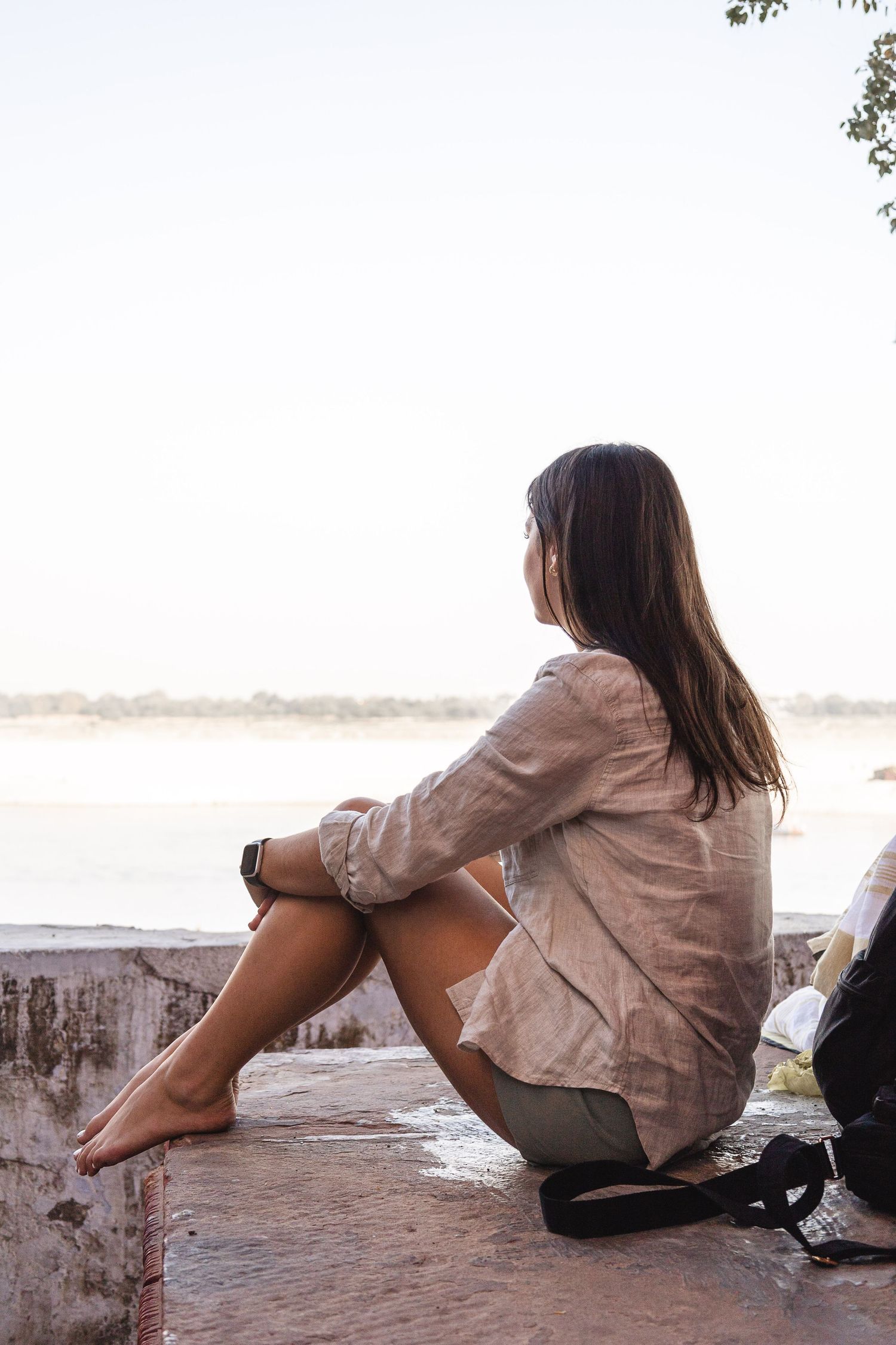 Young woman sitting barefoot on a stone wall gazing at a serene waterfront at sunset.