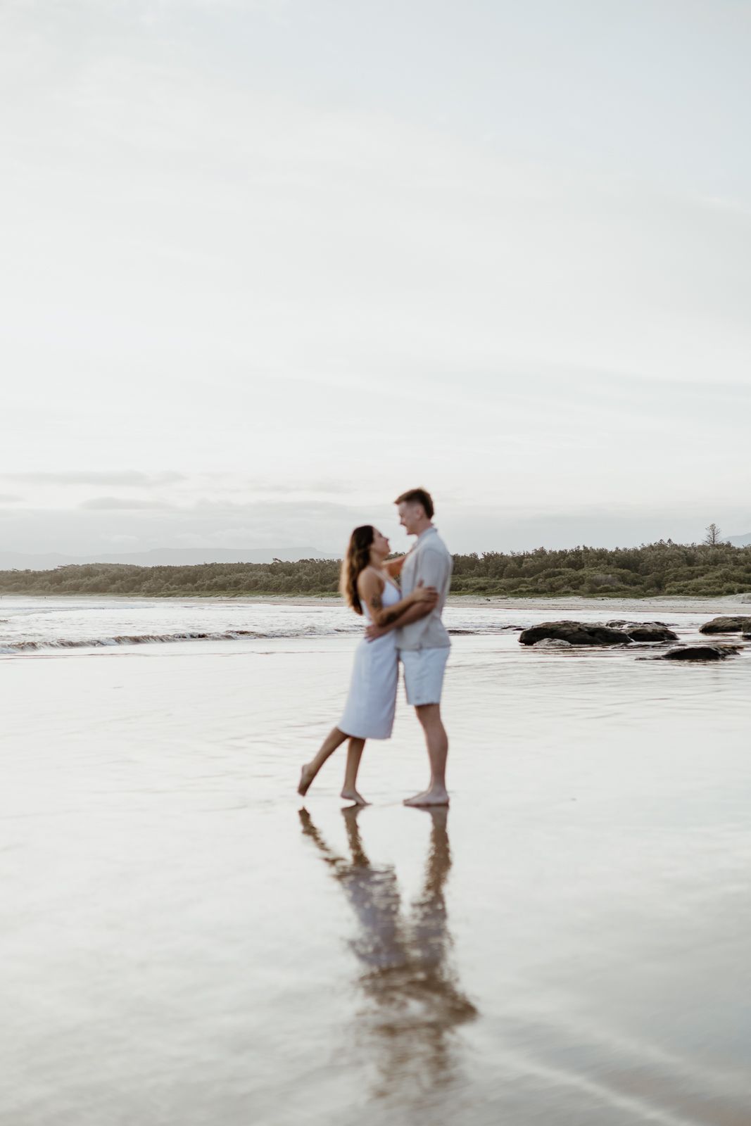 Couple embracing and gazing at each other on a calm beach with their reflection visible in the water.