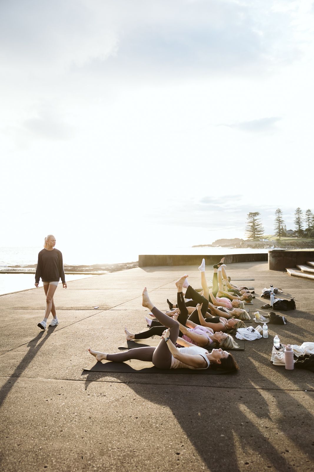 Fitness instructor walks past group lying on mats during outdoor seaside exercise class.