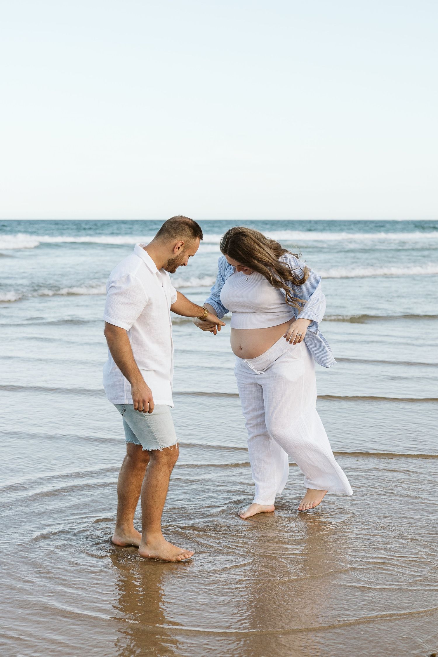 Couple holding hands on wet sandy beach shoreline, pregnant woman in white pants and blue shirt.